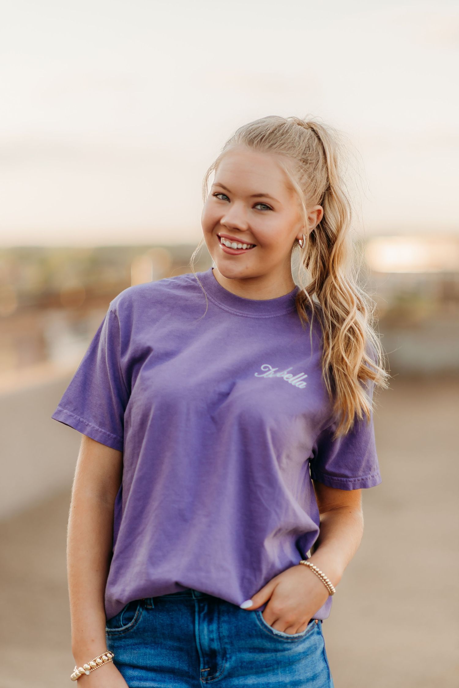 A casual portrait of someone wearing a purple t-shirt and blue jeans smiling outdoors at sunset.