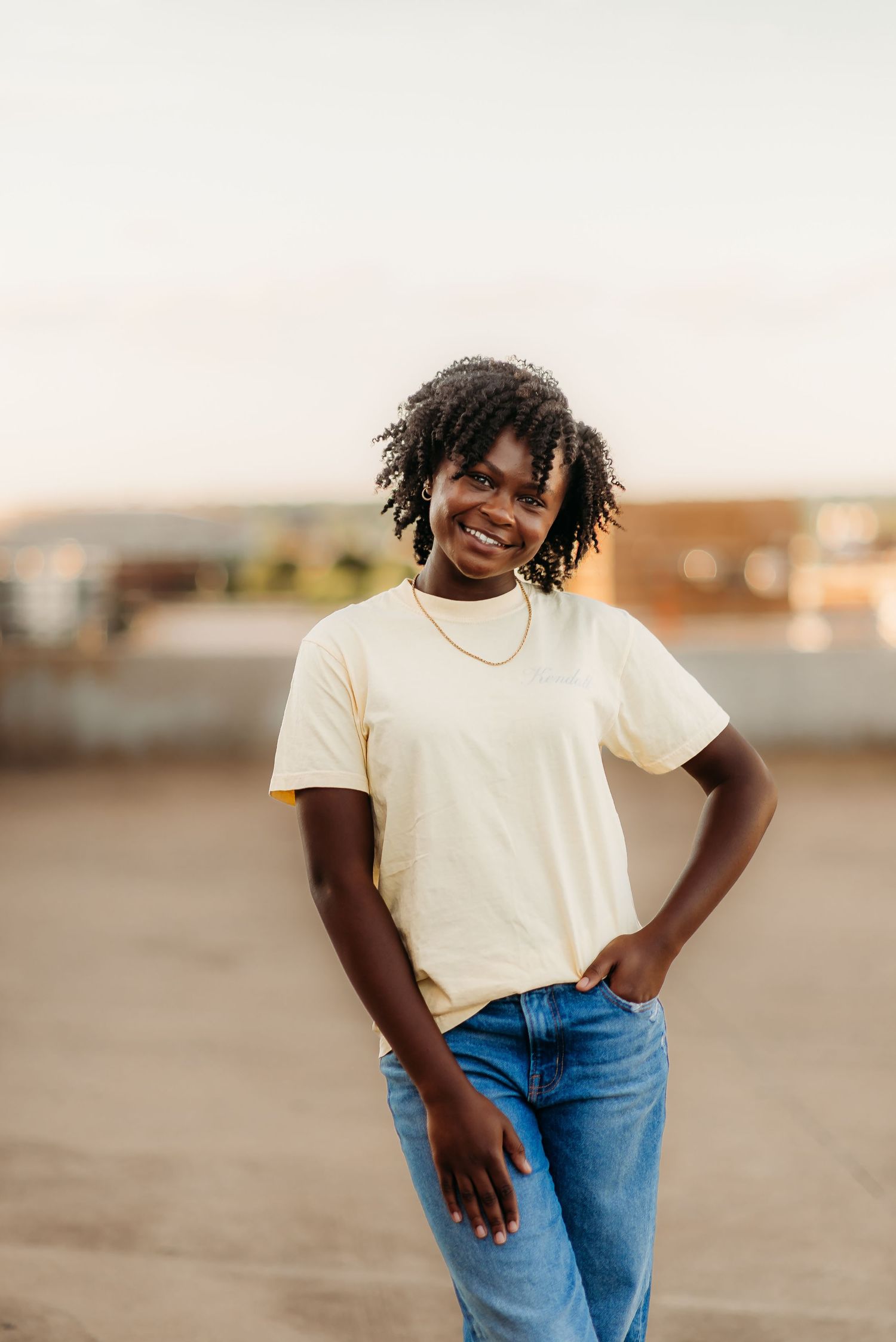 Person in casual white t-shirt and blue jeans poses outdoors with a joyful expression during golden hour.
