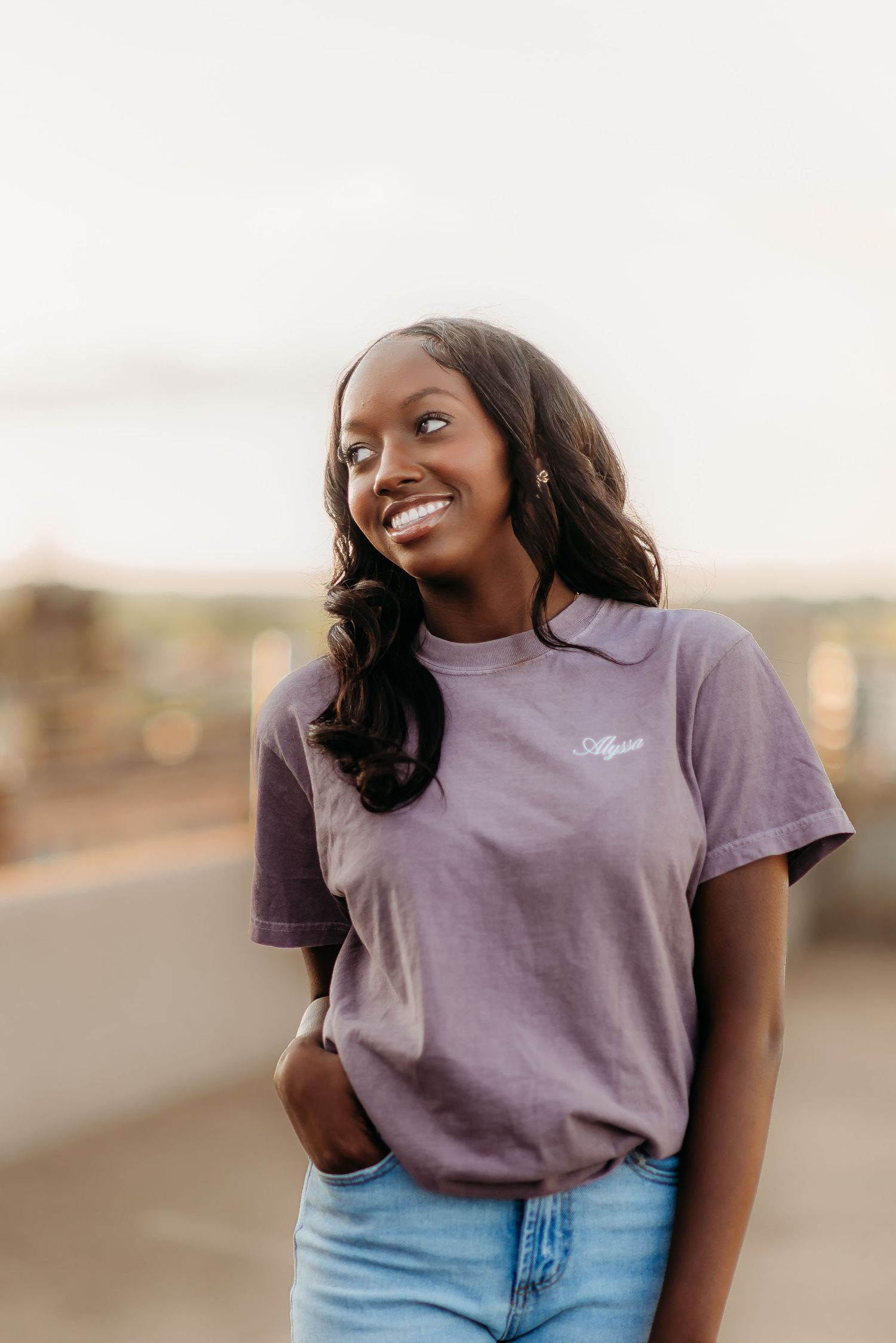 Woman in purple t-shirt and jeans smiling and posing against blurry outdoor backdrop during golden hour.