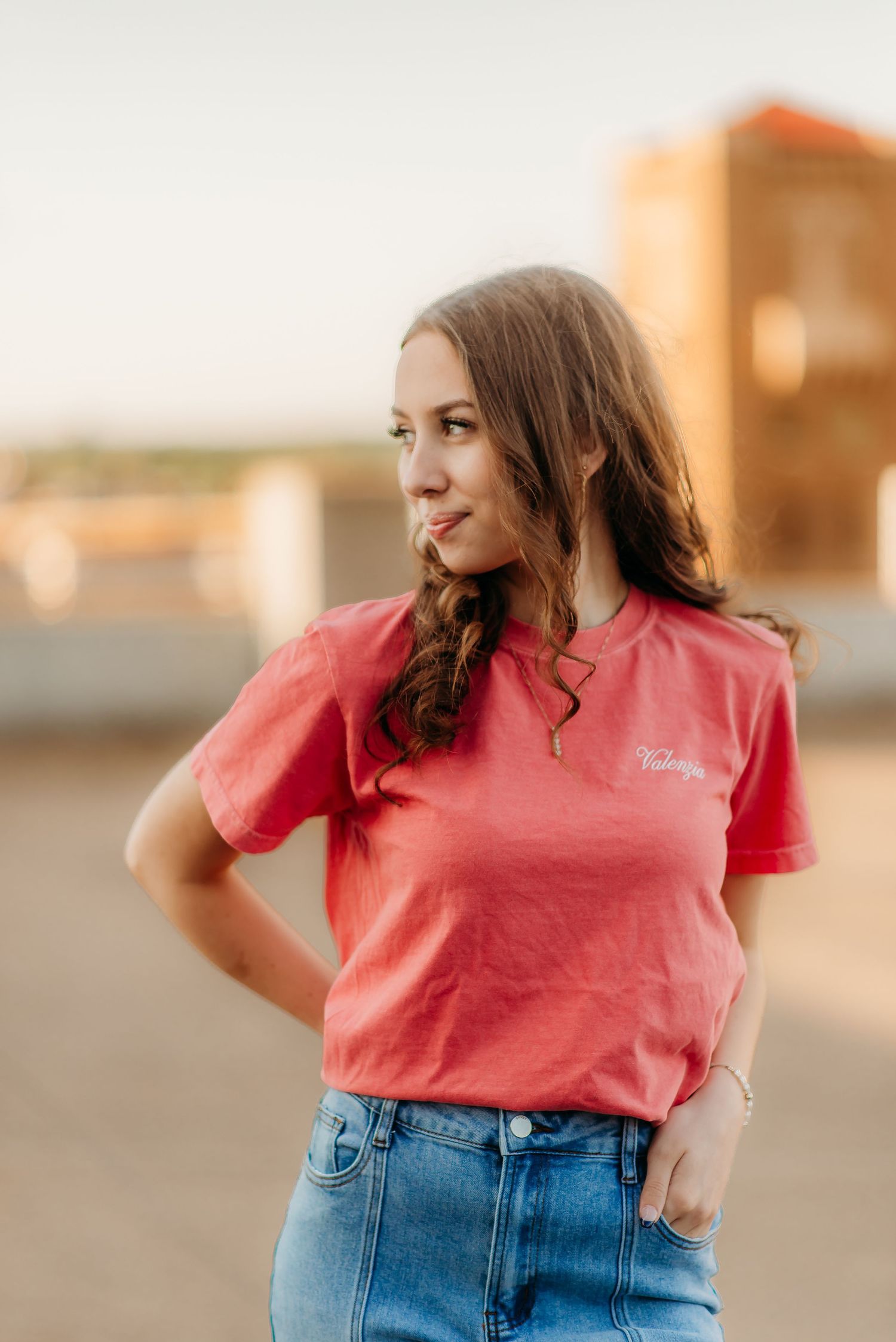 A woman in a coral t-shirt and denim skirt poses outdoors at golden hour with a contemplative gaze to the side.