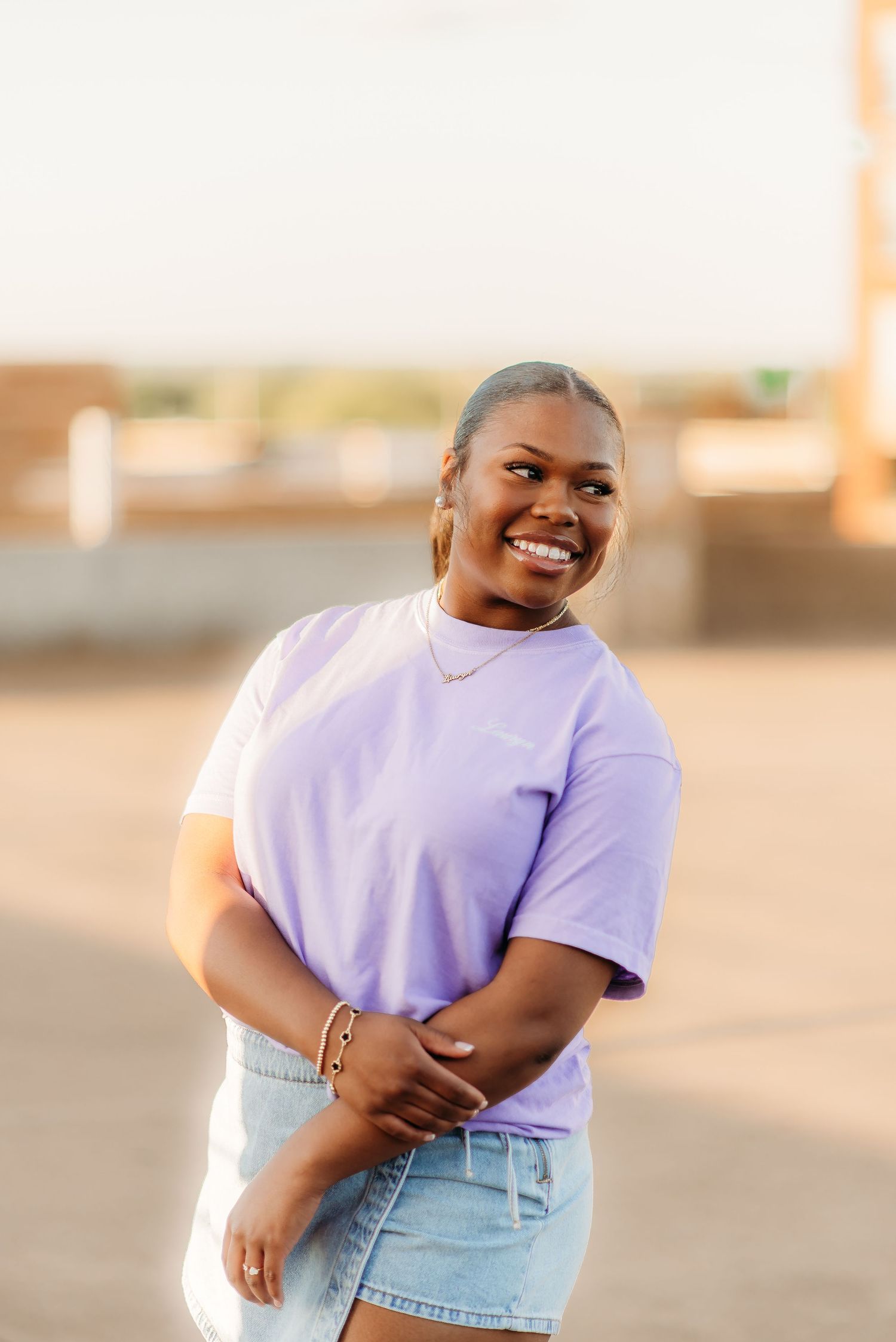 Person wearing a light purple t-shirt and denim shorts poses joyfully against a warm sunset background outdoors.