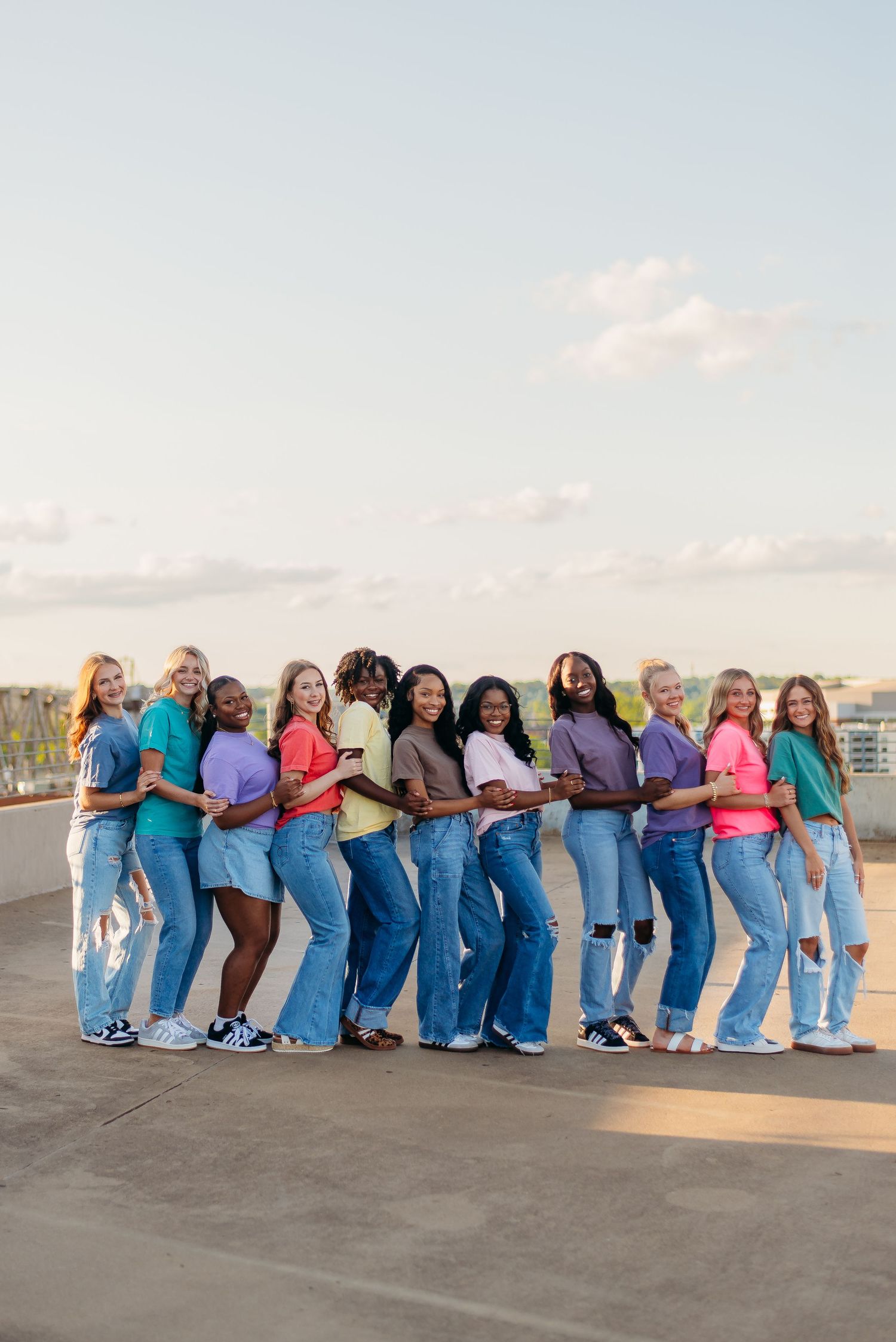 A group of people wearing colorful tops and jeans pose together in a line on a rooftop at sunset.
