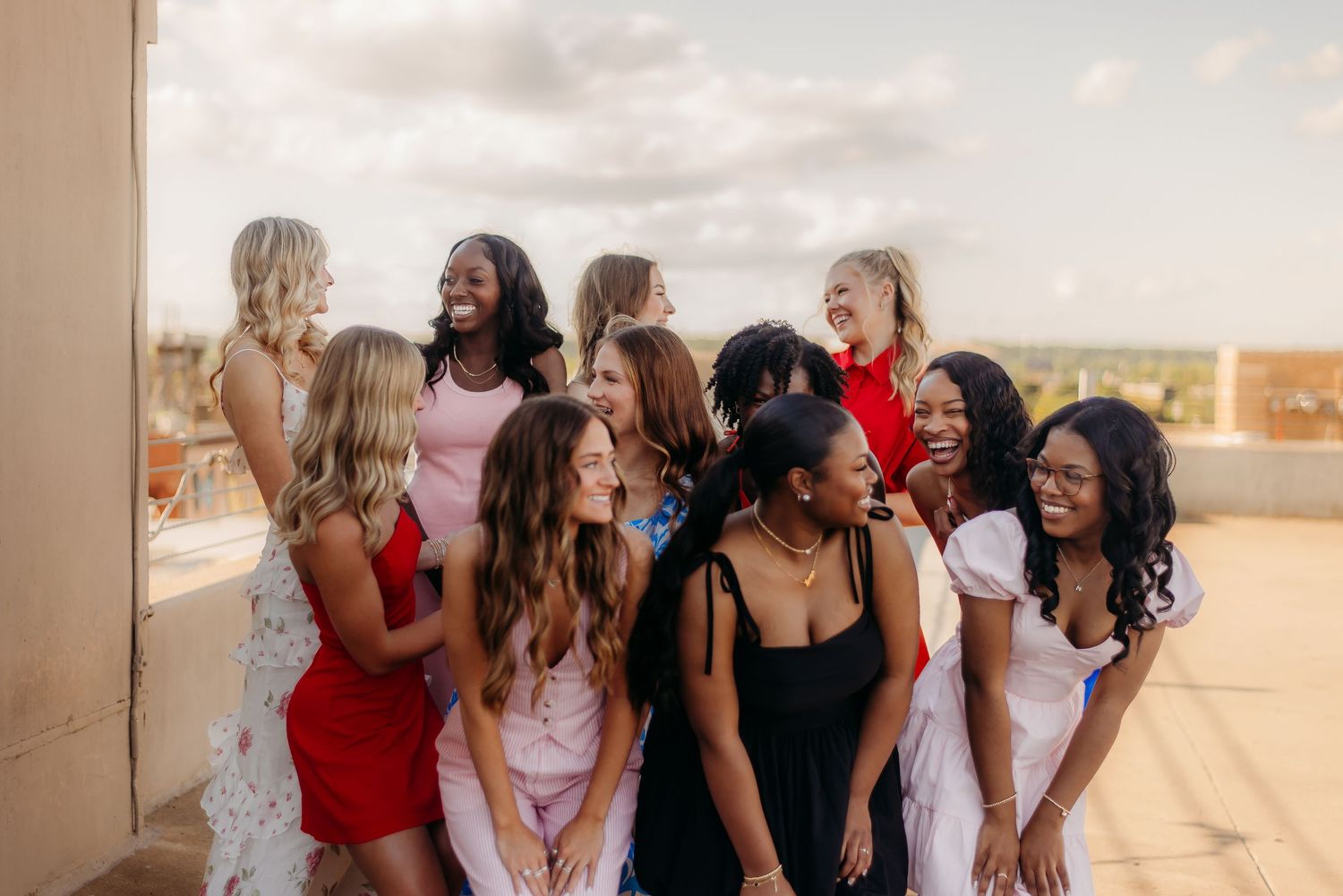 A diverse group of friends in colorful dresses laughing and socializing together on a rooftop during sunset.