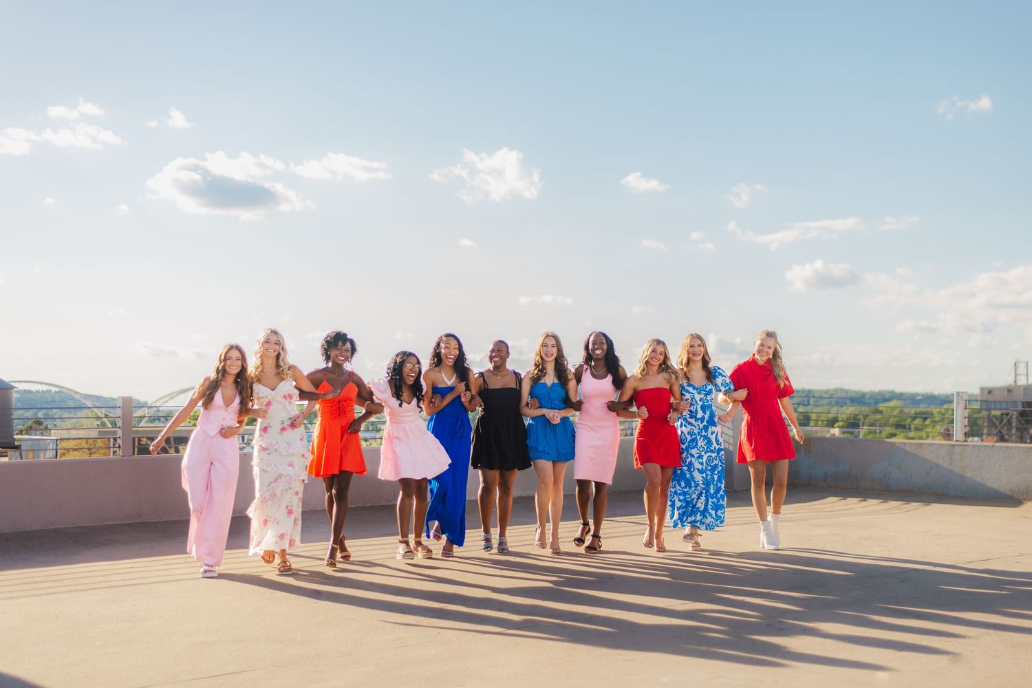 A group in colorful dresses stands together on a rooftop patio with a scenic view on a sunny day.