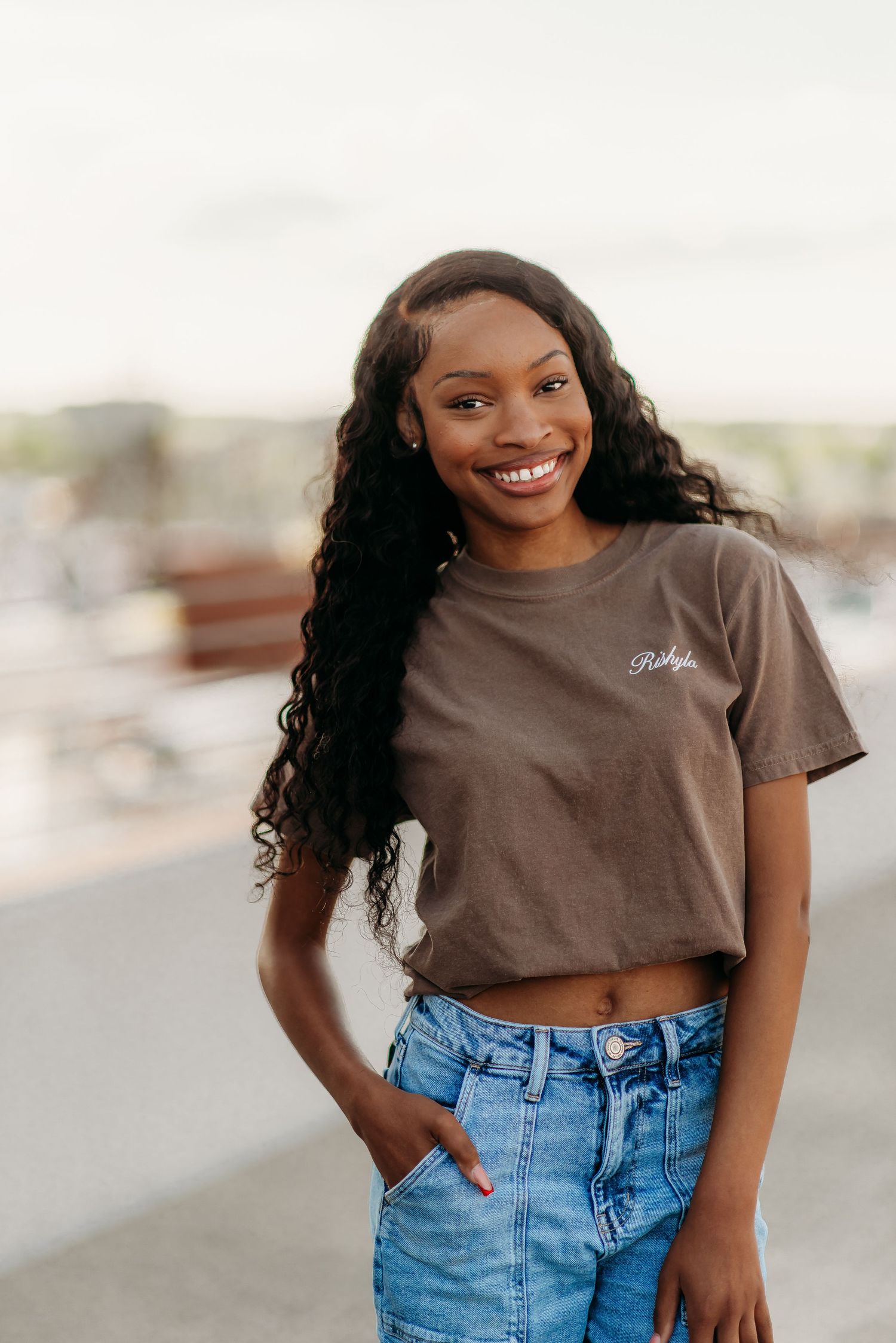 Smiling person wearing brown crop top t-shirt and blue jeans poses outdoors with long curly dark hair.