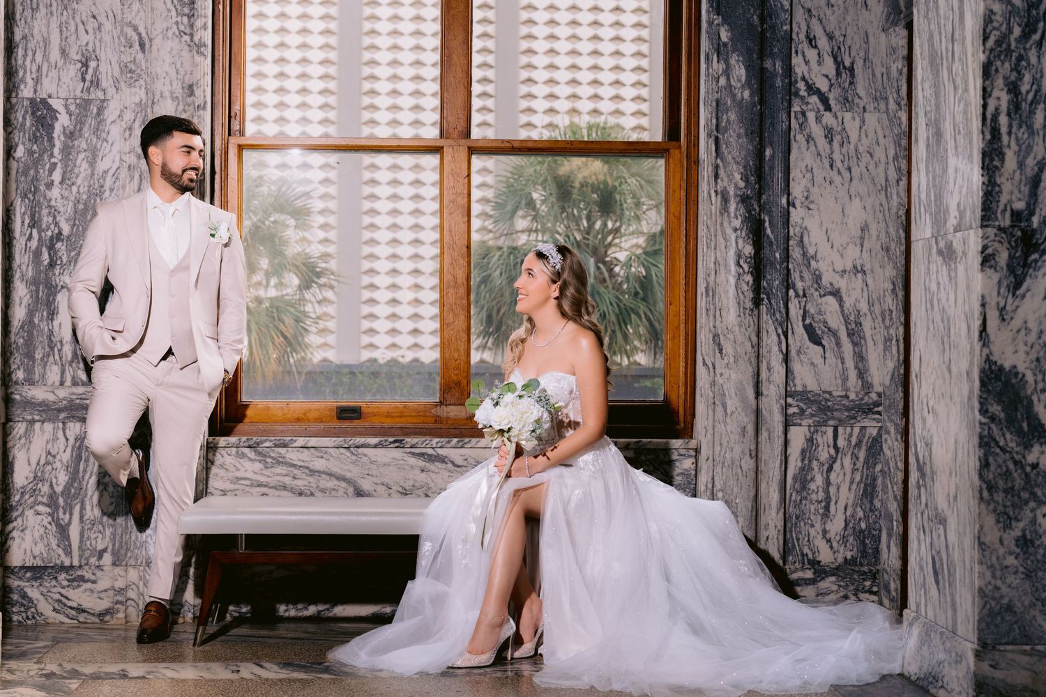 A wedding couple poses elegantly by a window in a marble-walled room at Le Meridien Tampa, with the bride seated in a white gown.
