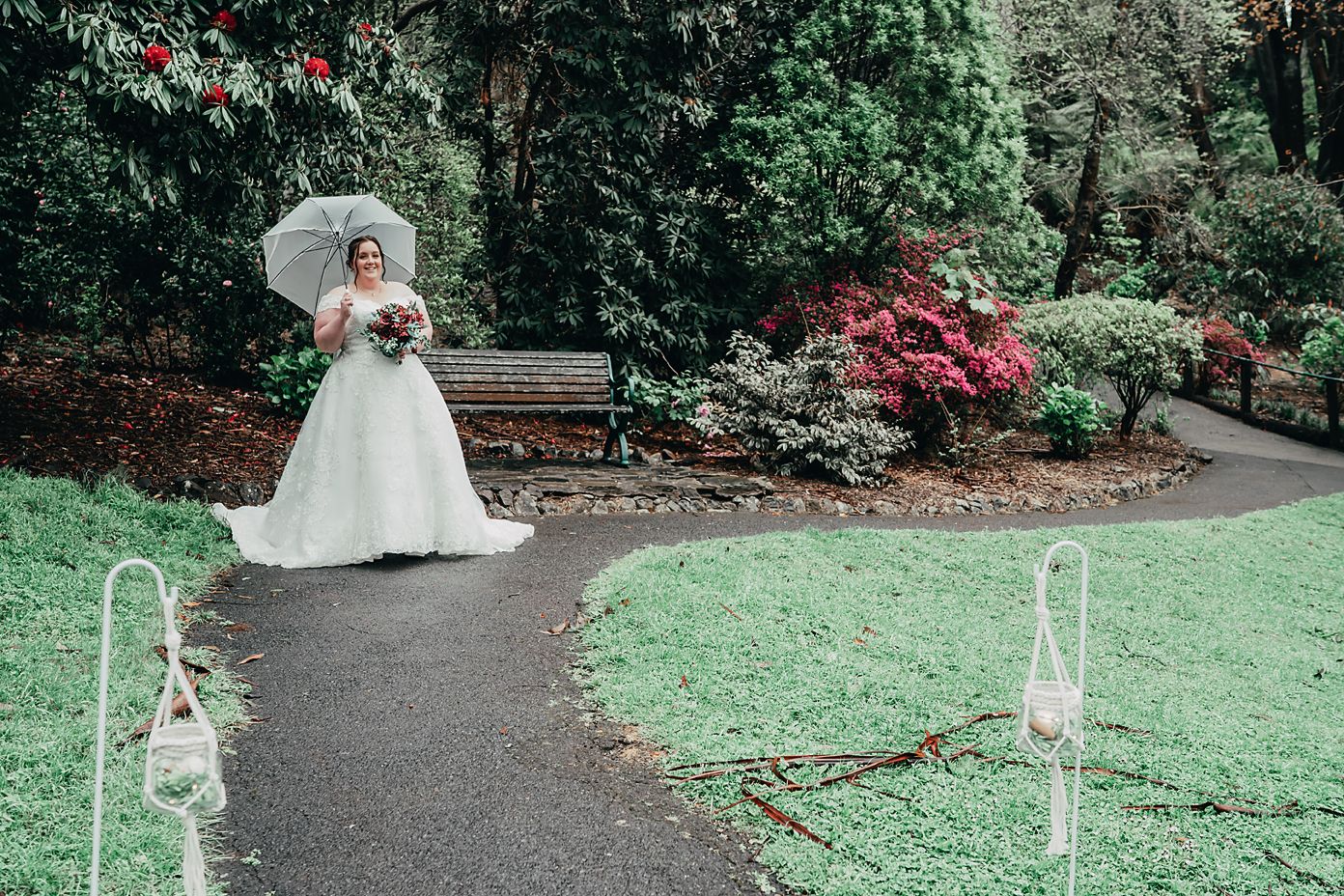 Bride in white wedding dress walking along winding garden path between decorative white chairs and flowering bushes.