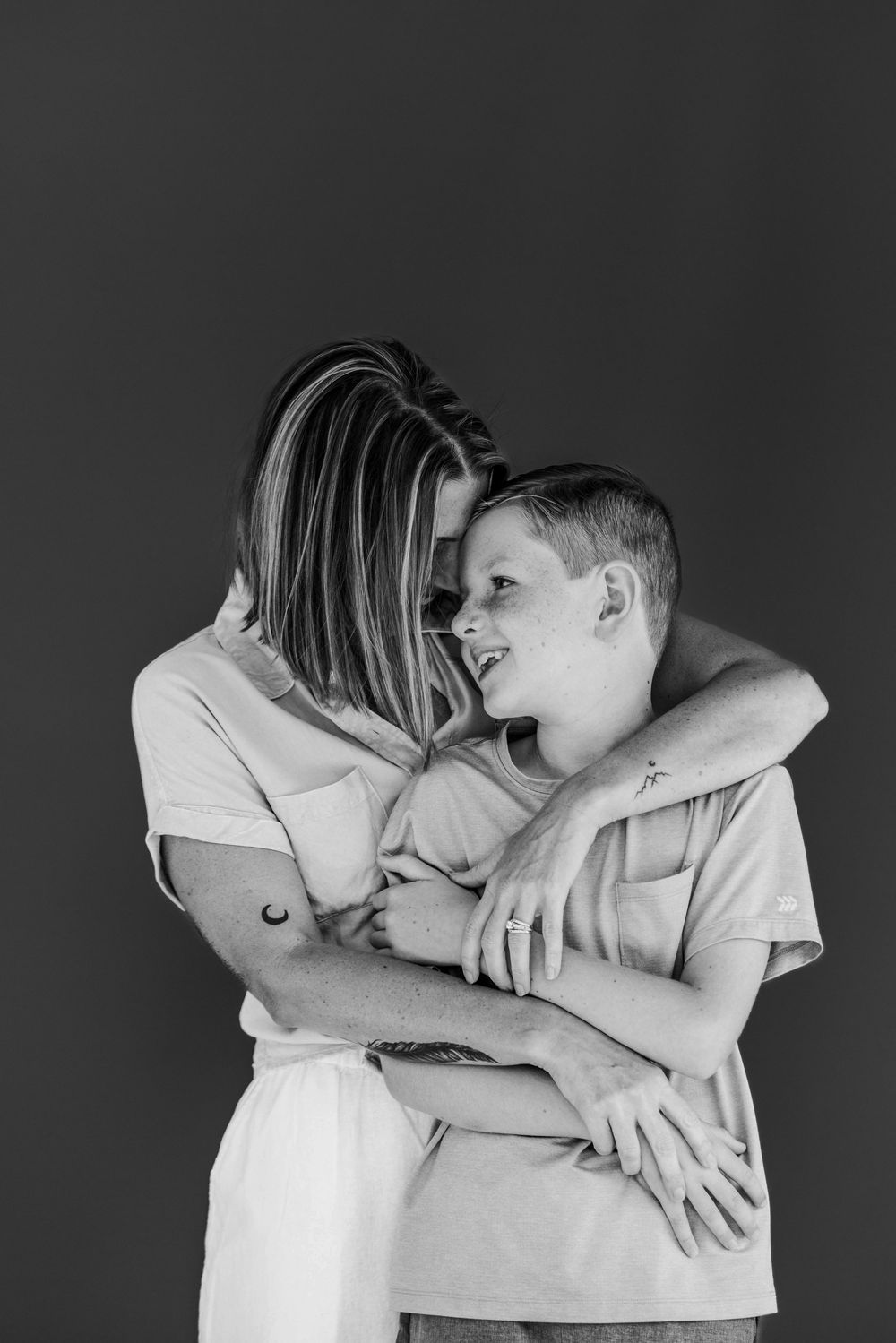 A tender black and white portrait of a mother and son embracing and smiling against a dark background.