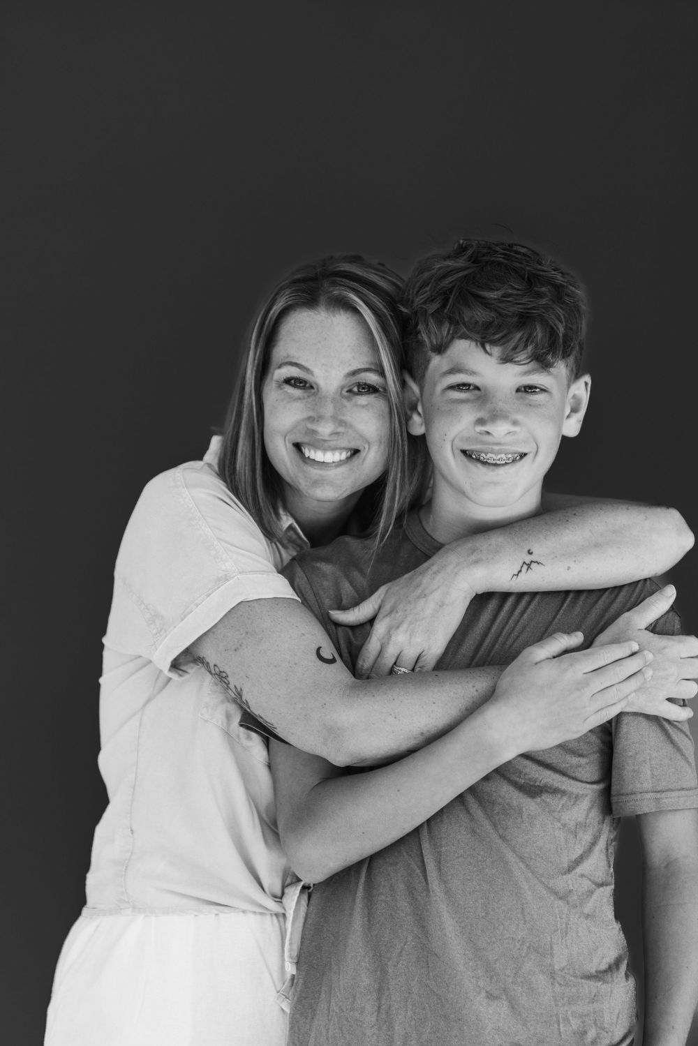 Black and white portrait of a mother and teenage son smiling and embracing against a dark background.