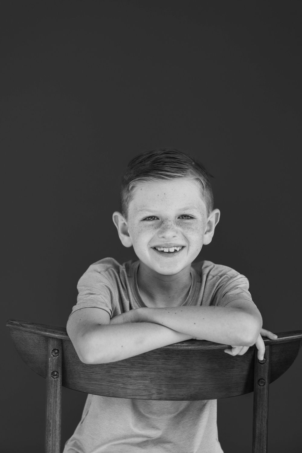 Black and white portrait of a child resting arms on a wooden chair back with a bright smile.