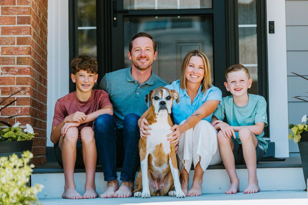 A family sits together with their brown and white dog on their front porch steps on a sunny day.