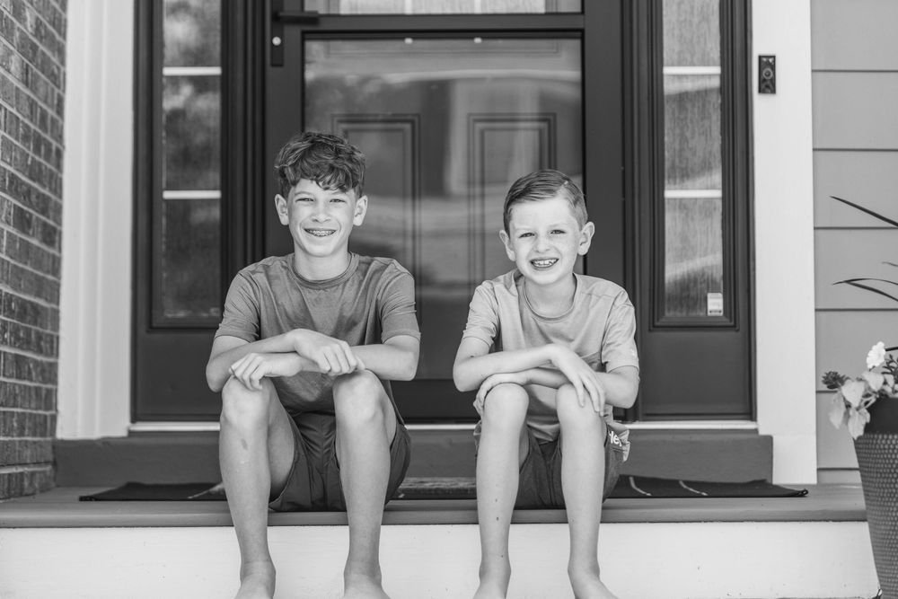 Two children sit together smiling on the front steps of a house in black and white.