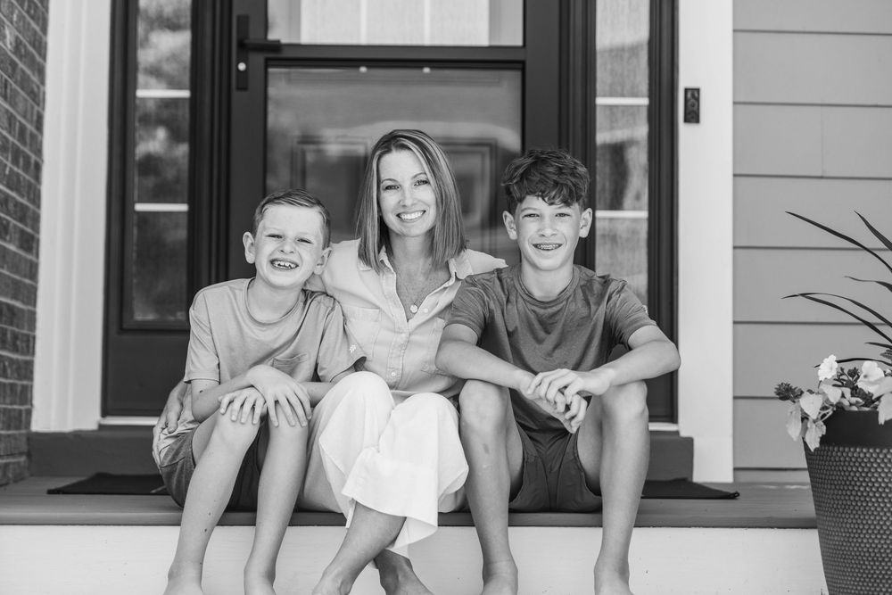 A candid family portrait in black and white shows three people sitting together on the front steps of their home.