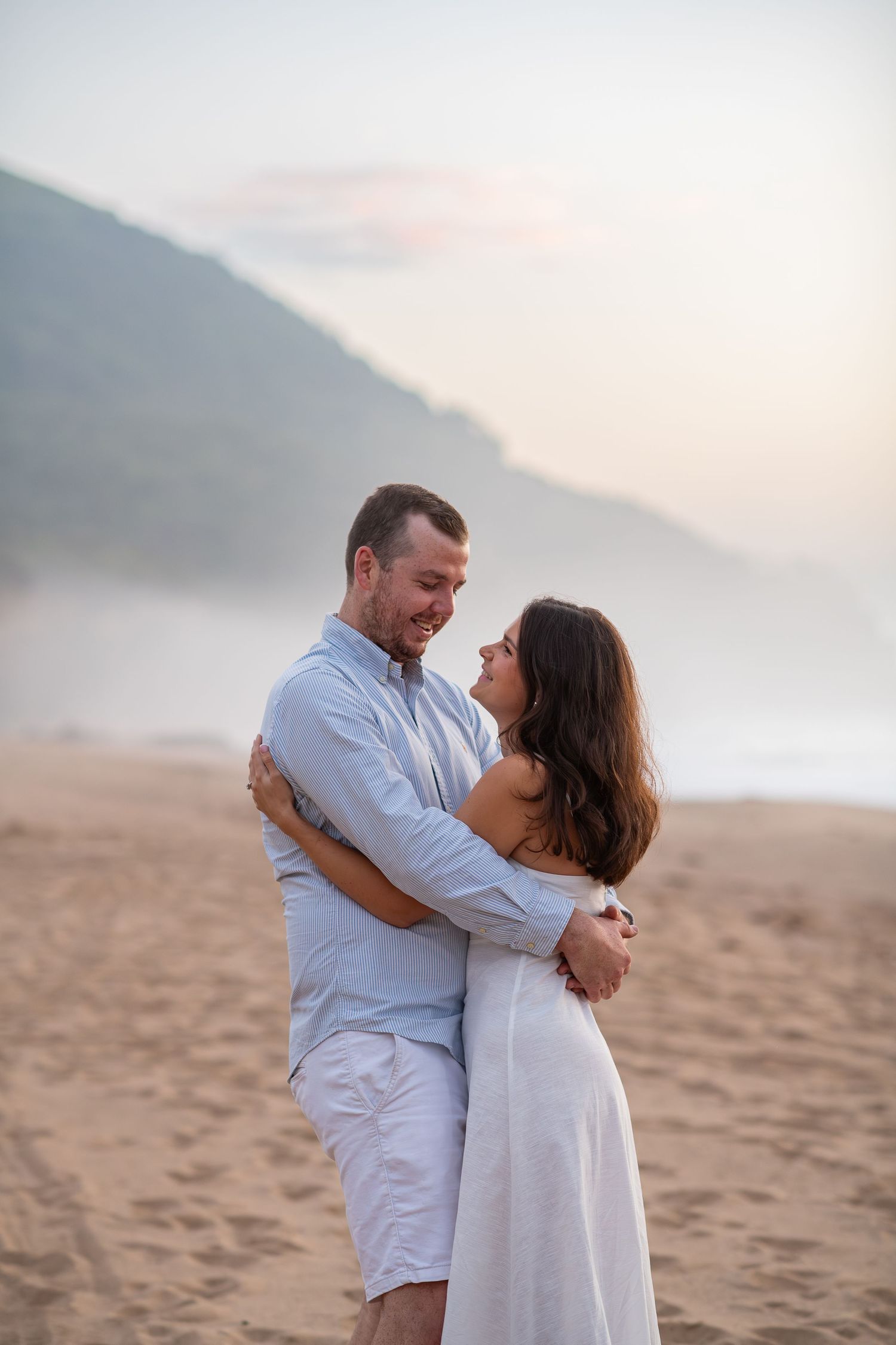 A couple shares a romantic moment on a misty beach at sunset wearing white outfits.