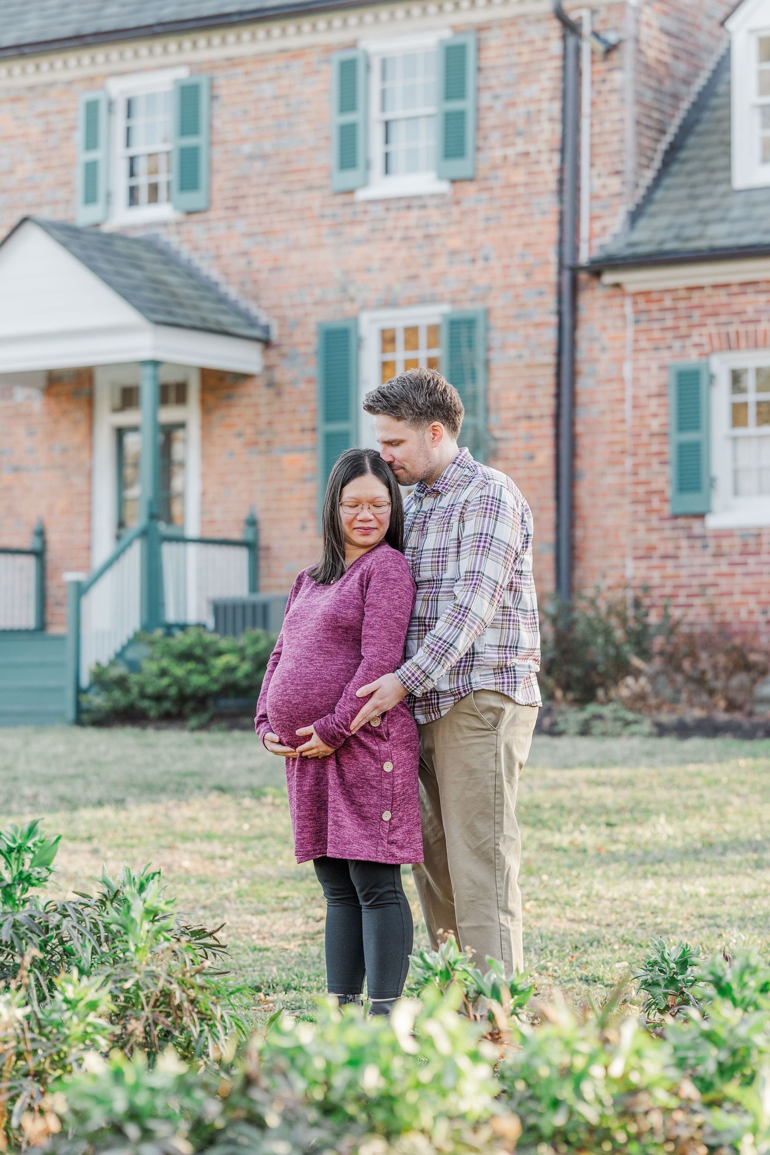 A couple stands together in a garden outside a brick house with green shutters.