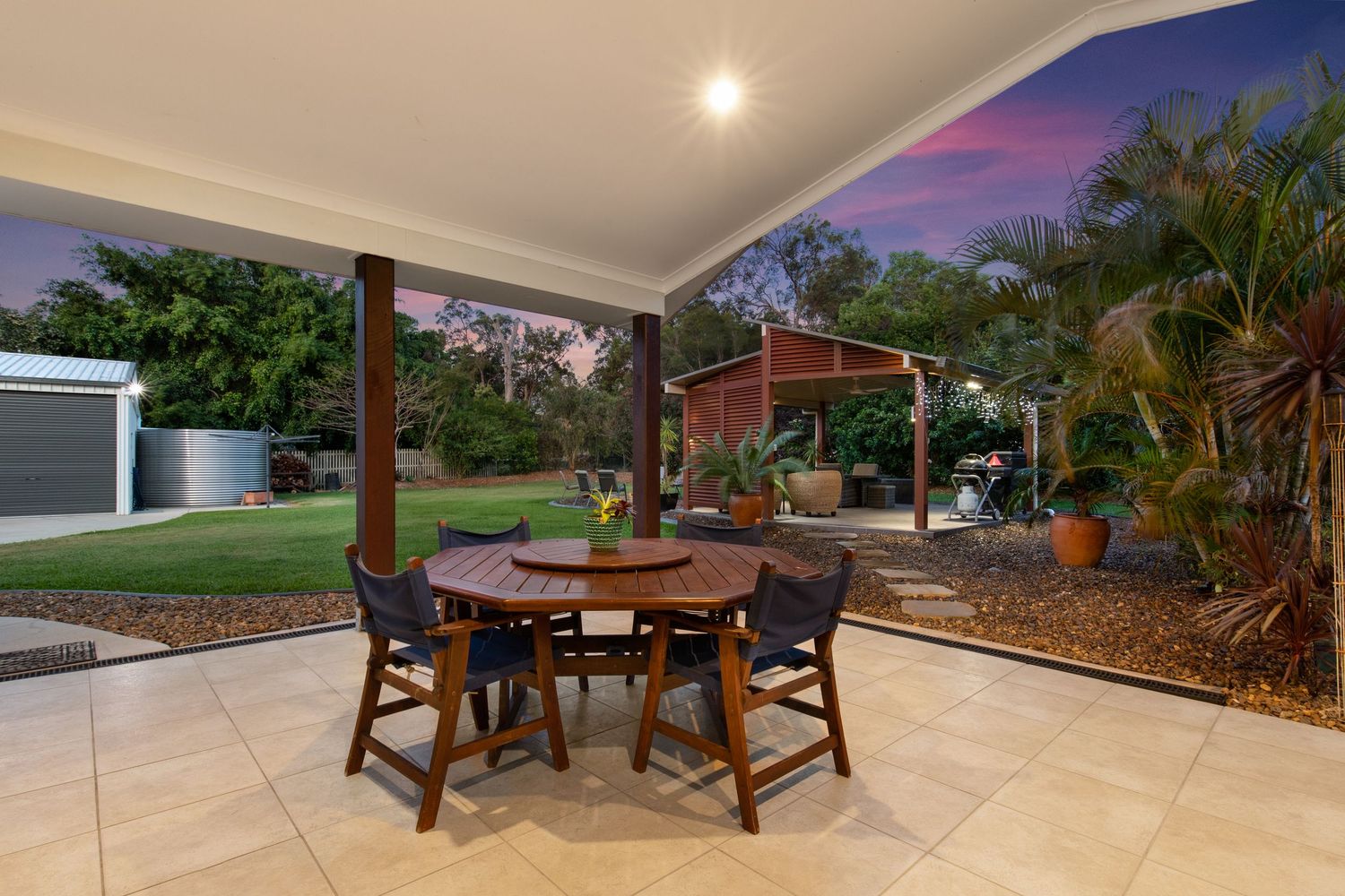 Covered alfresco dining area with timber table and chairs overlooking a large landscaped backyard at dusk.