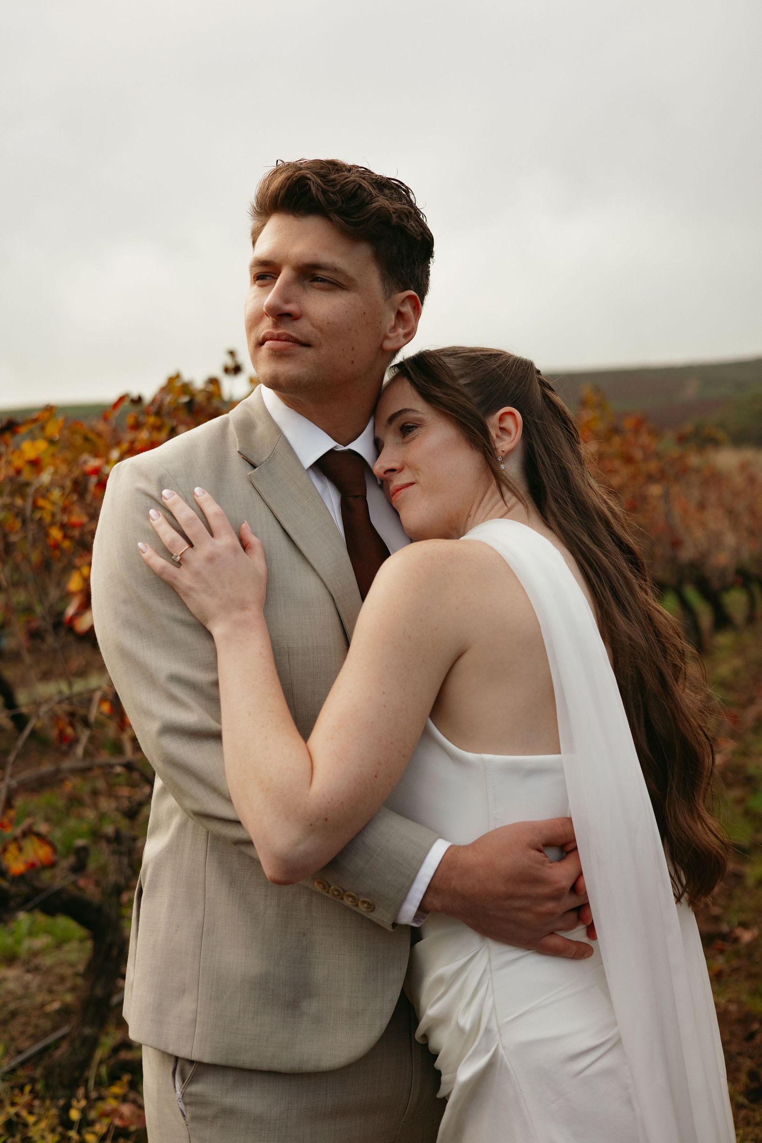 Bride and groom embracing in an autumn vineyard, groom in beige suit with burgundy tie.