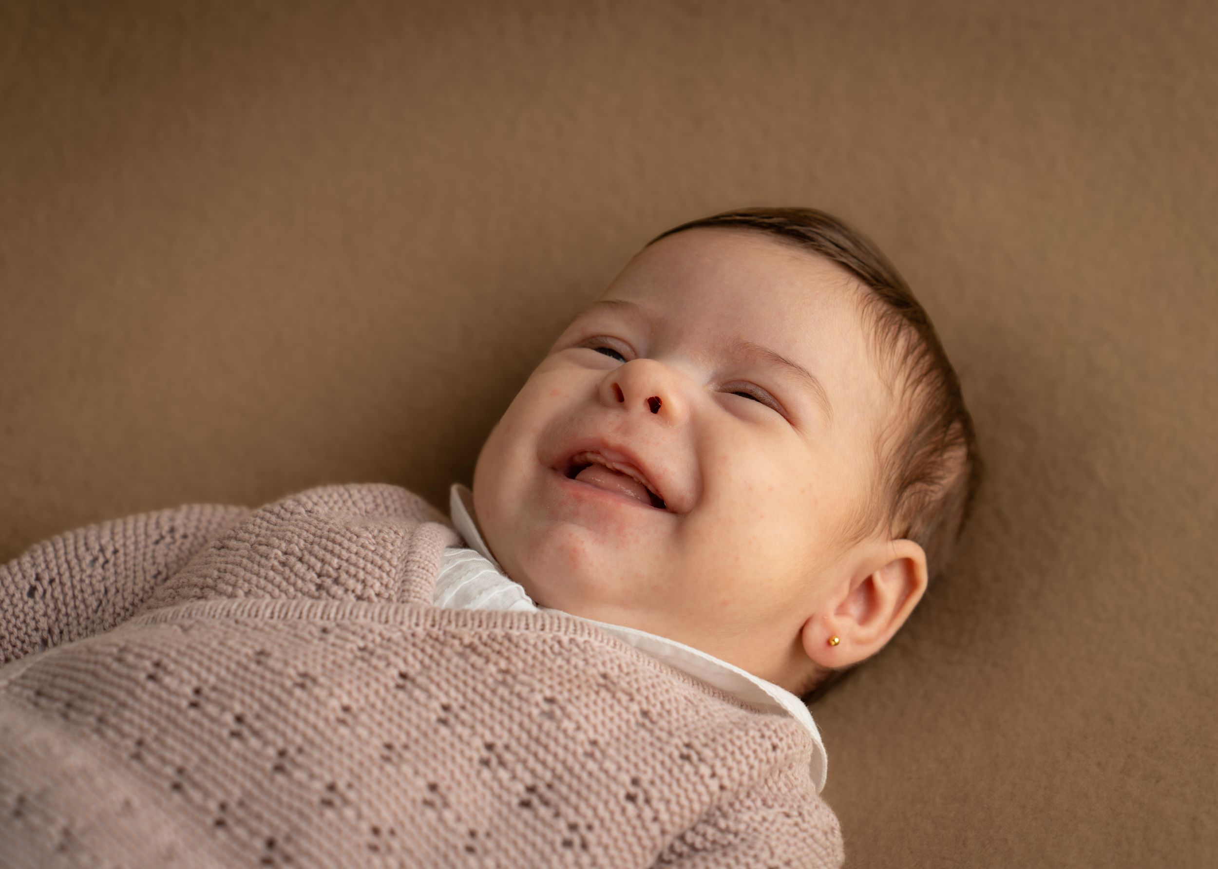 A smiling newborn baby wrapped in a soft pink knit blanket lying on a warm beige background.
