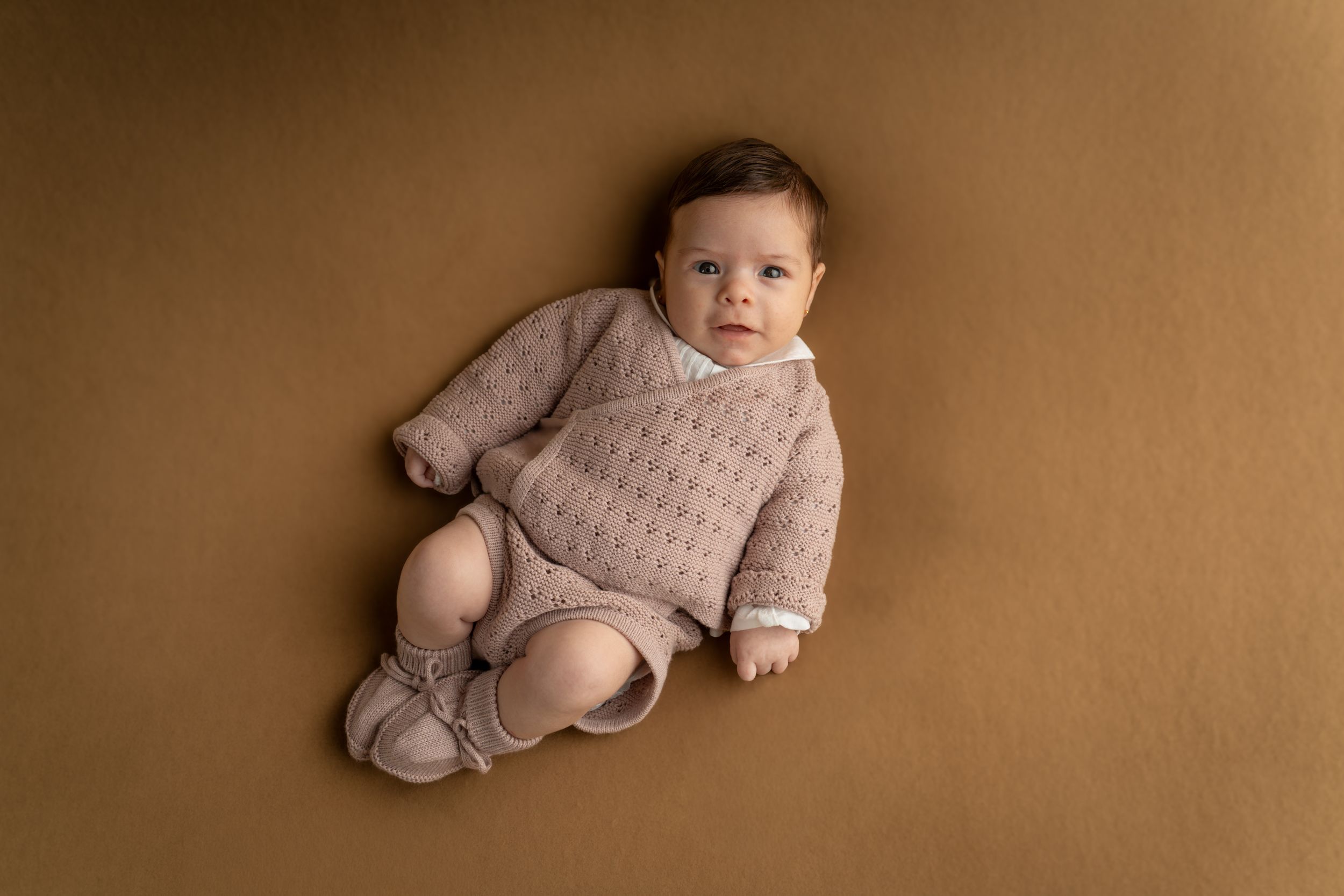 An adorable baby in a beige knit romper lying on a brown backdrop gazing at the camera.