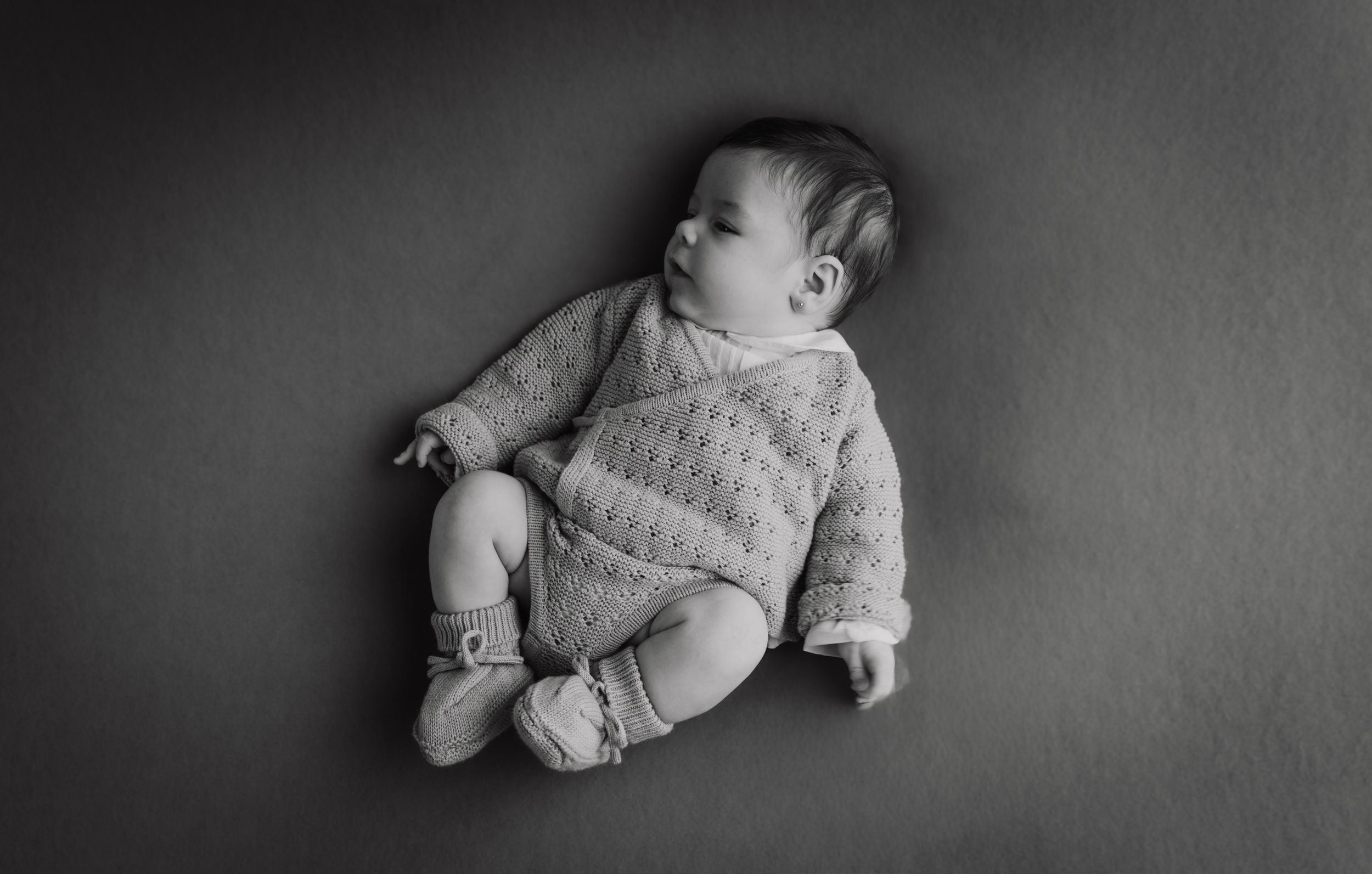 Black and white photo of a newborn baby in a knit romper lying on a dark background.