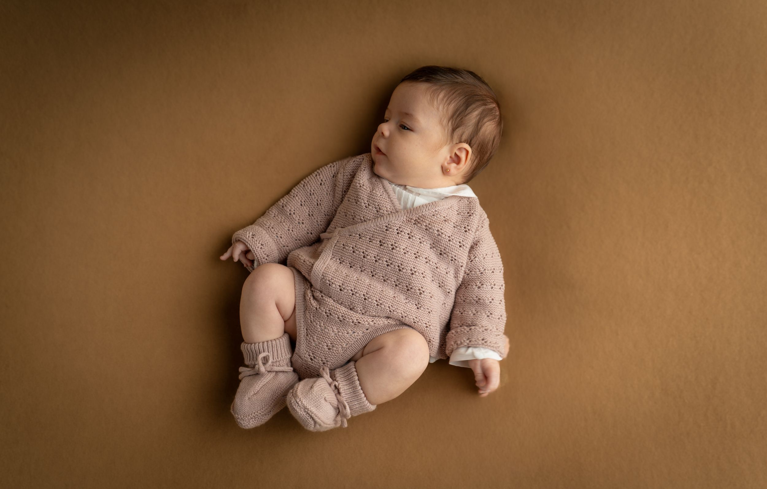 Newborn baby in a pink knit romper and booties lying on a warm brown background.