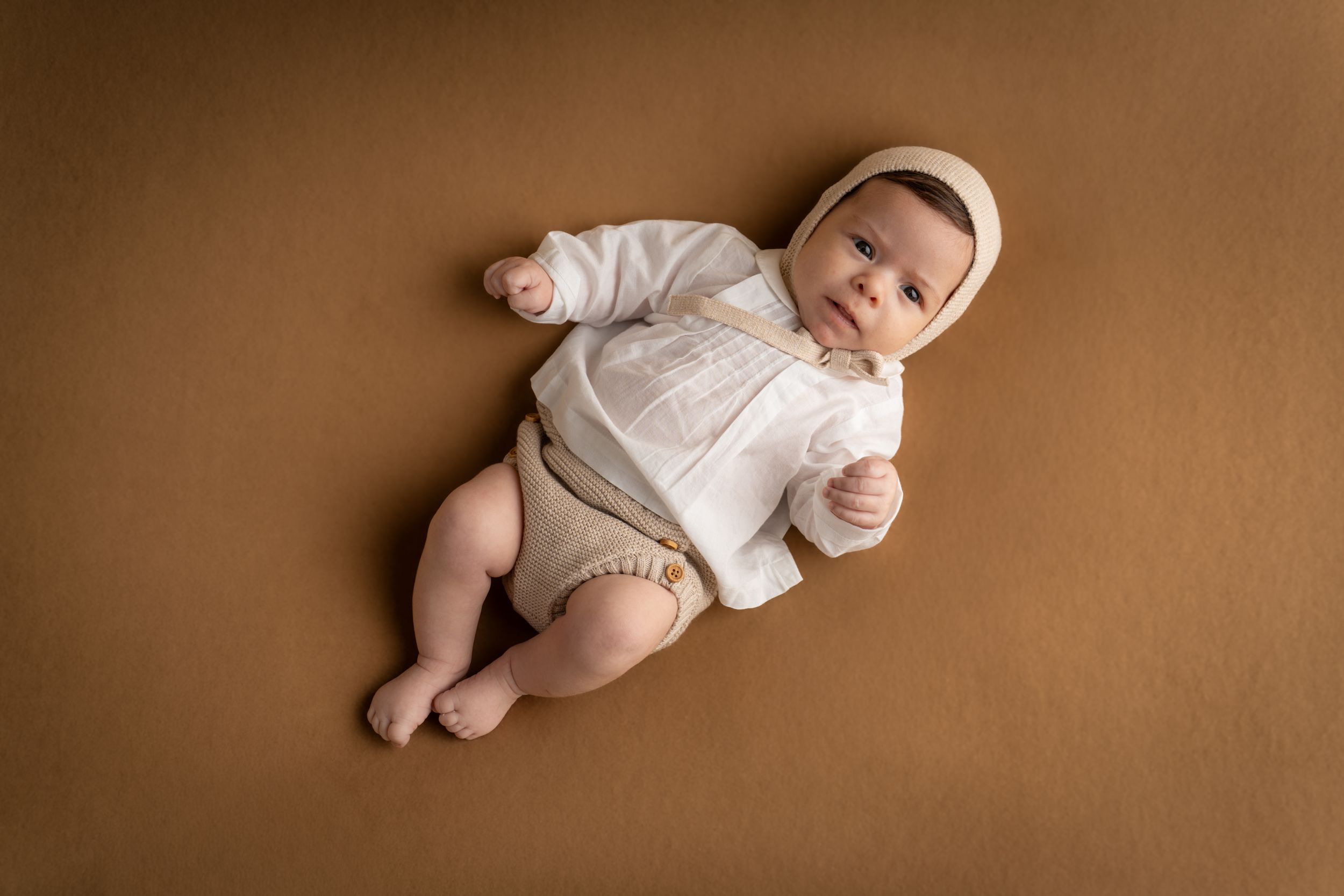 Adorable baby in white shirt and bonnet lying on a brown backdrop looking up.