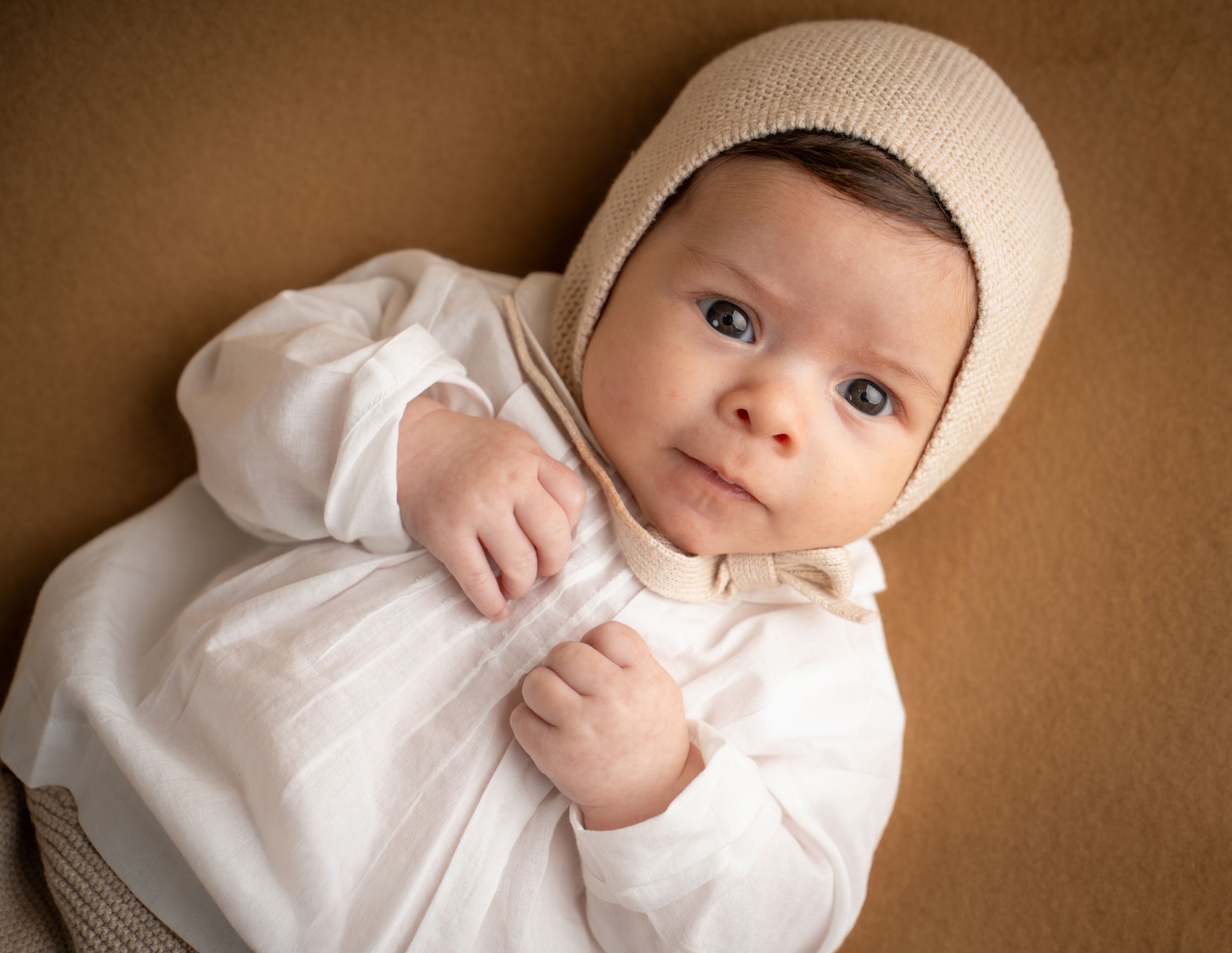 Close-up portrait of a newborn baby in a white outfit and beige knit bonnet.