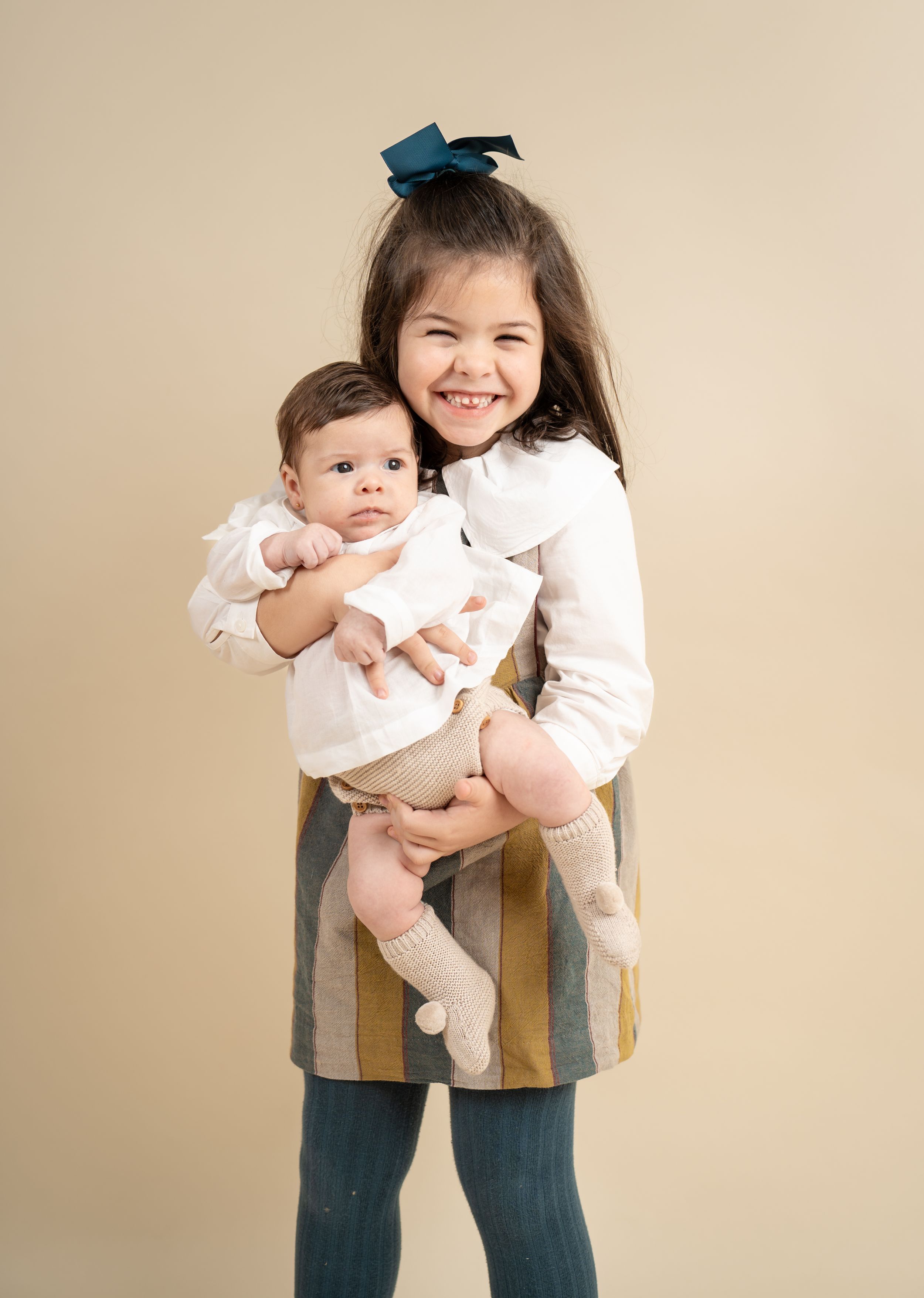 Smiling little girl with teal bow holding newborn baby sibling in studio portrait.