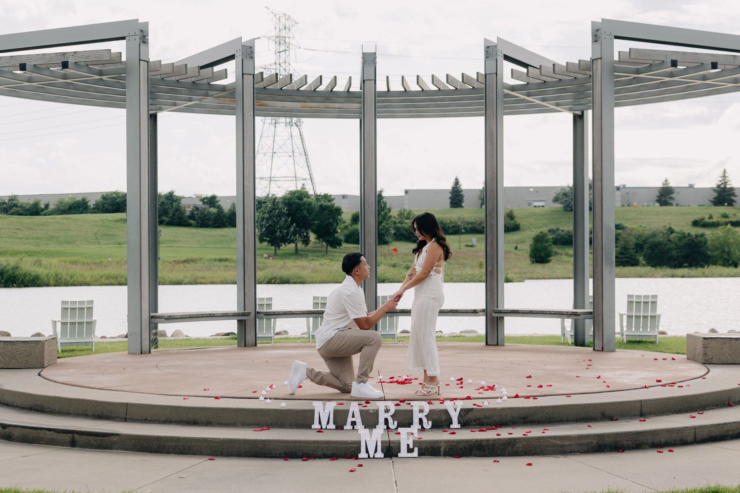 A romantic proposal scene at a circular gazebo overlooking a lake with rose petals scattered on the ground.