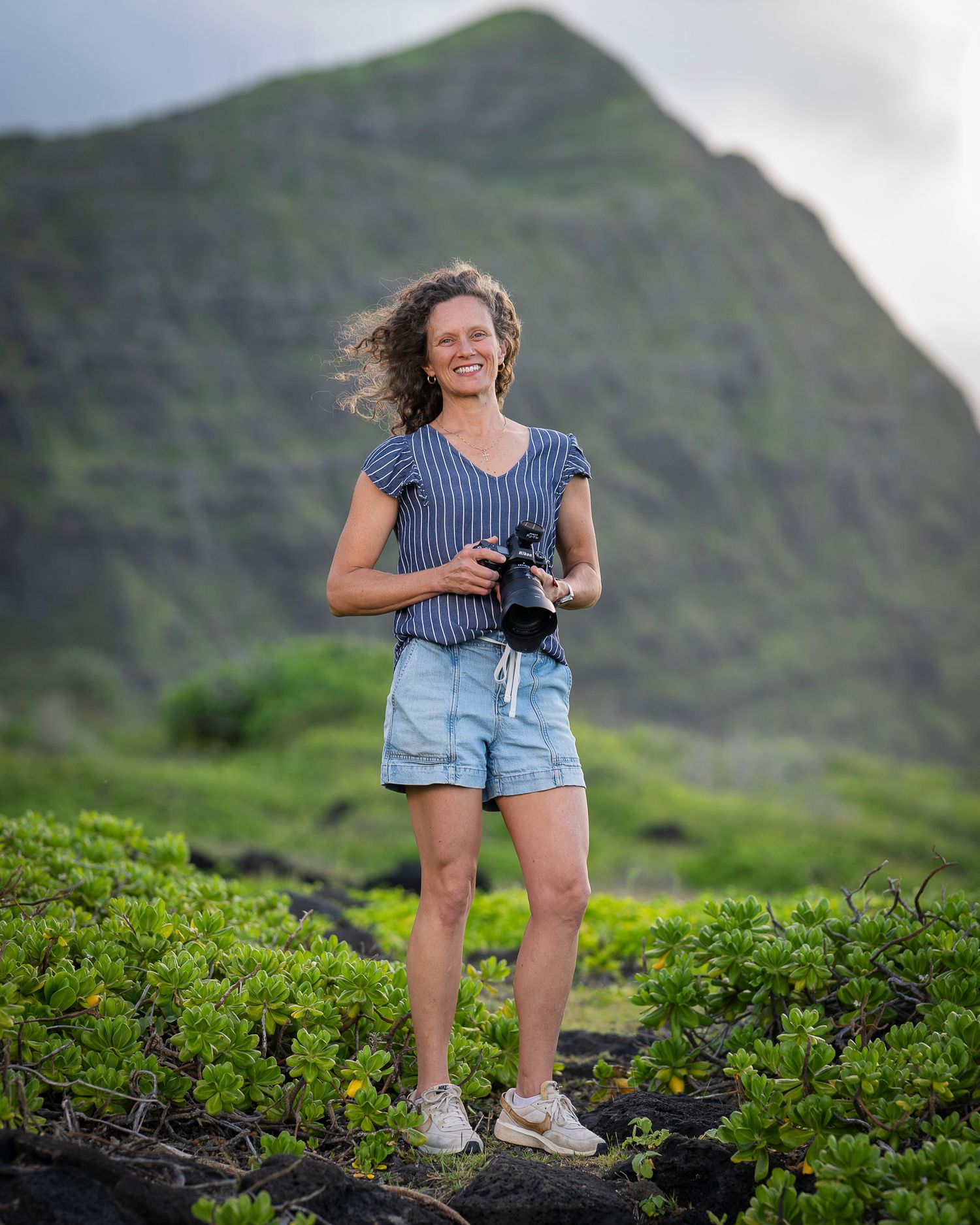 Jenna Ettlich Photography, a Honolulu Family Photographer stands with Nikon camera near Oahu mountains at Makapuu Beach in Hawaii