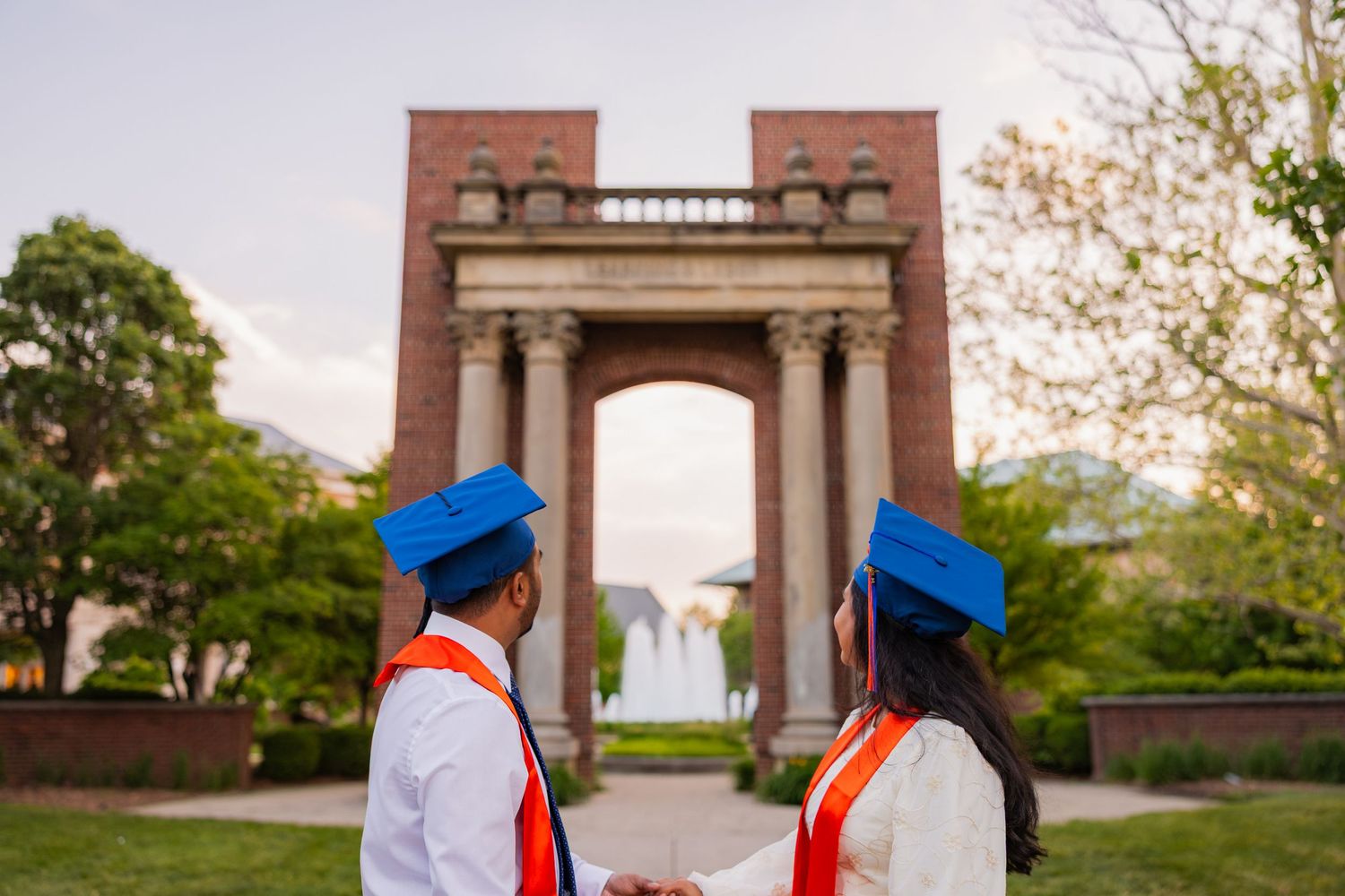 UIUC graduate couple with graduation attire looking up at Hallene Gateway with a fountain in the background