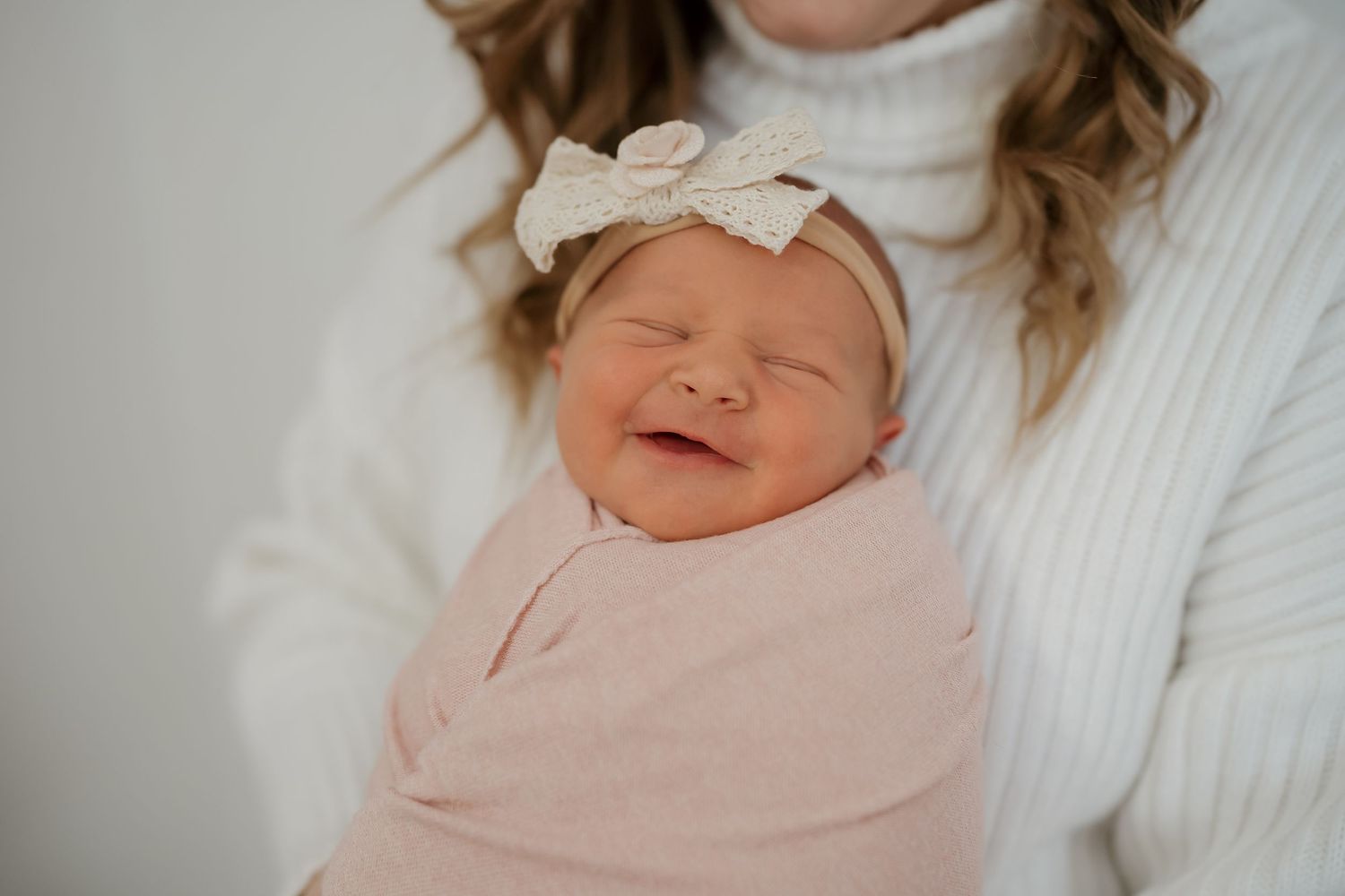 A sleeping newborn baby wrapped in a soft pink swaddle blanket rests peacefully on white bedding.