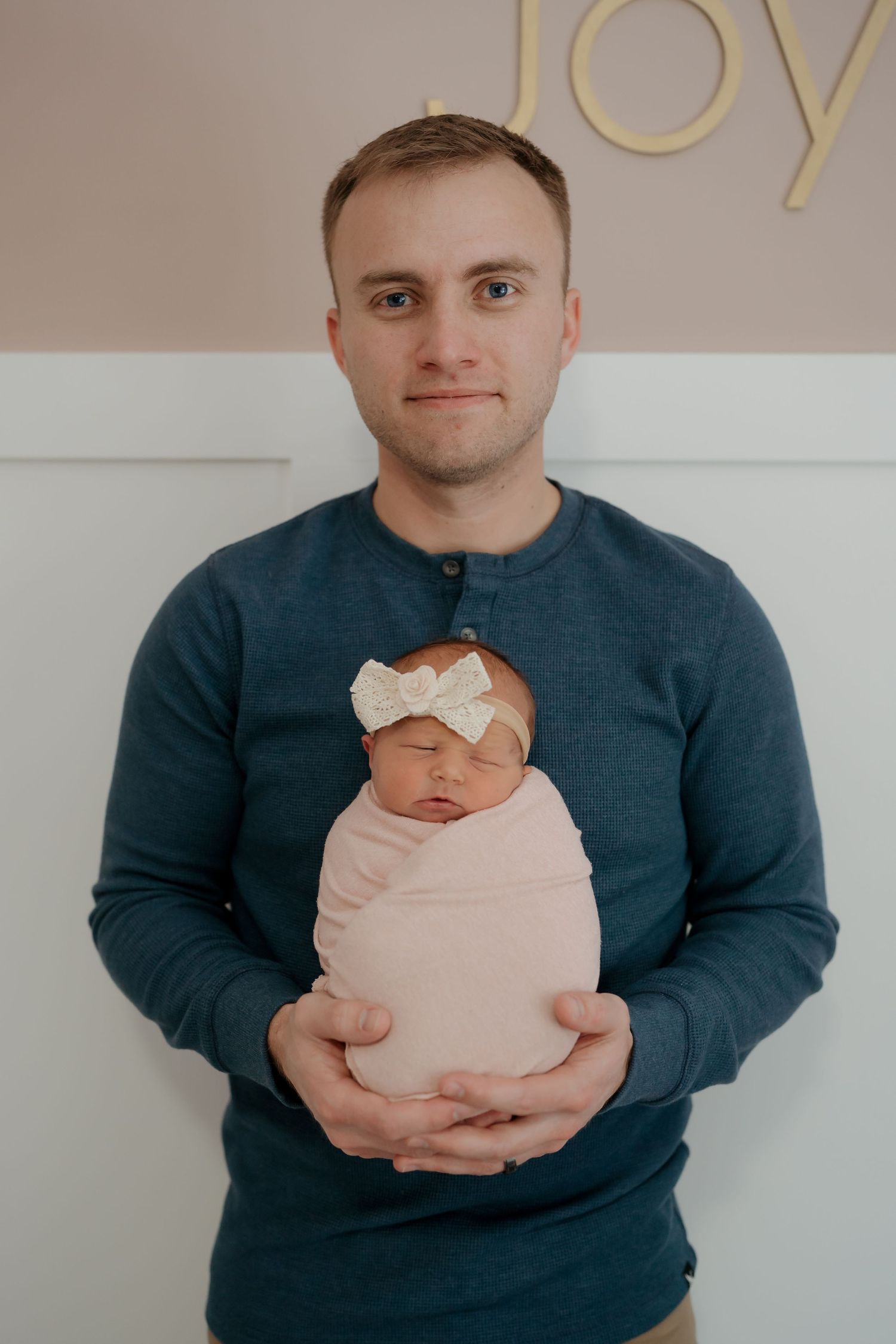 Parent in navy sweater holds sleeping newborn baby wearing pink outfit and white bow headband against neutral wall.