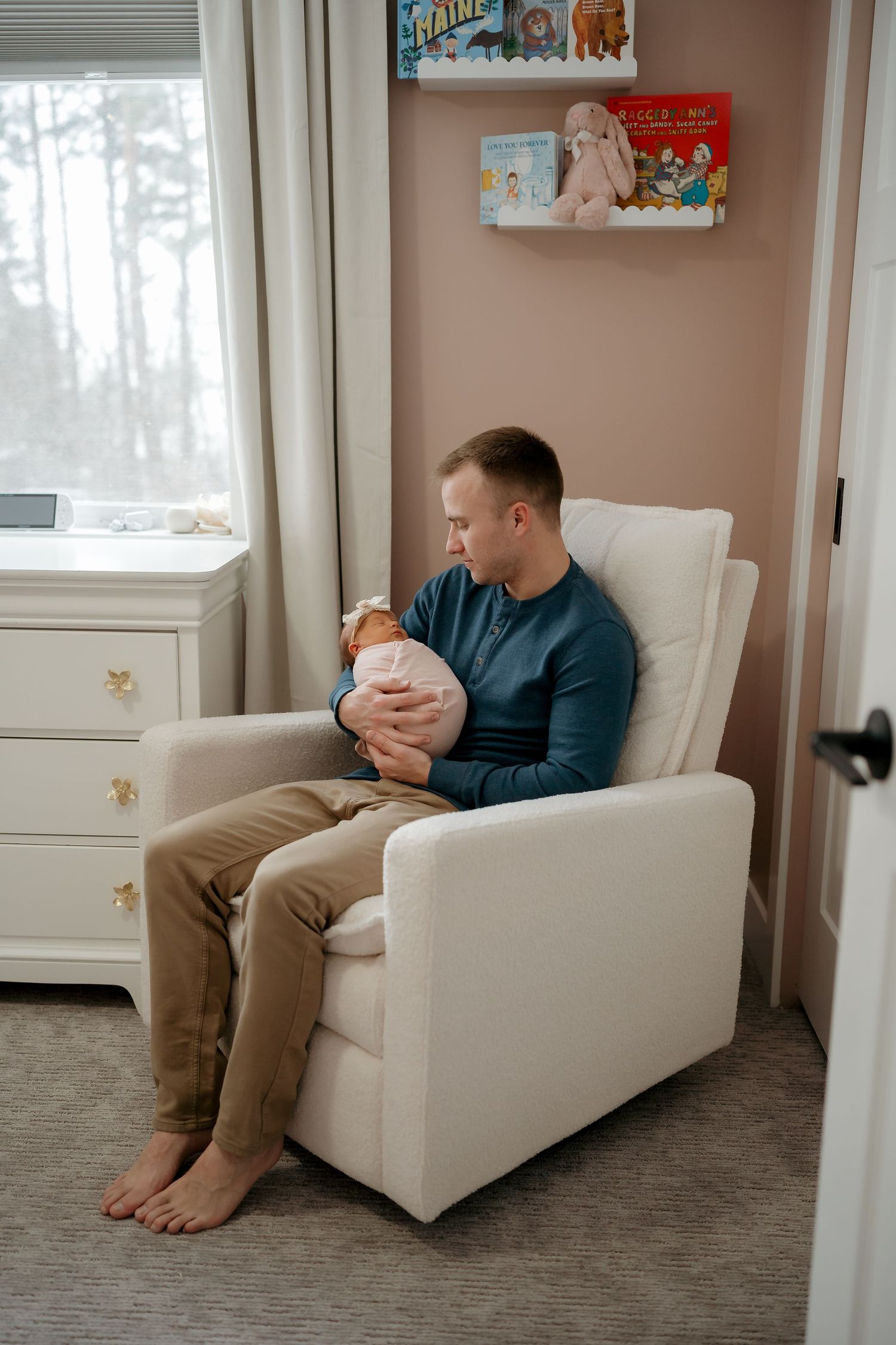 A parent sits barefoot in a white armchair holding a newborn baby in a cozy nursery with children's books on the wall.