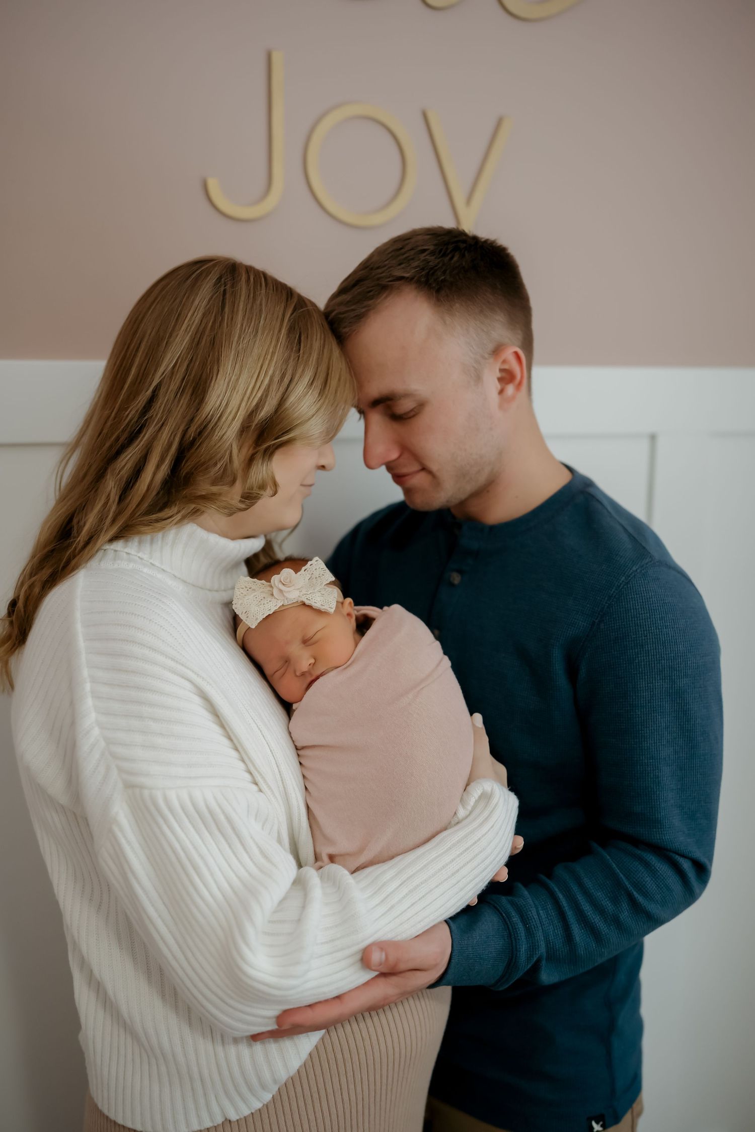 Parents in white and navy sweaters share tender moment with sleeping newborn in pink against neutral background.