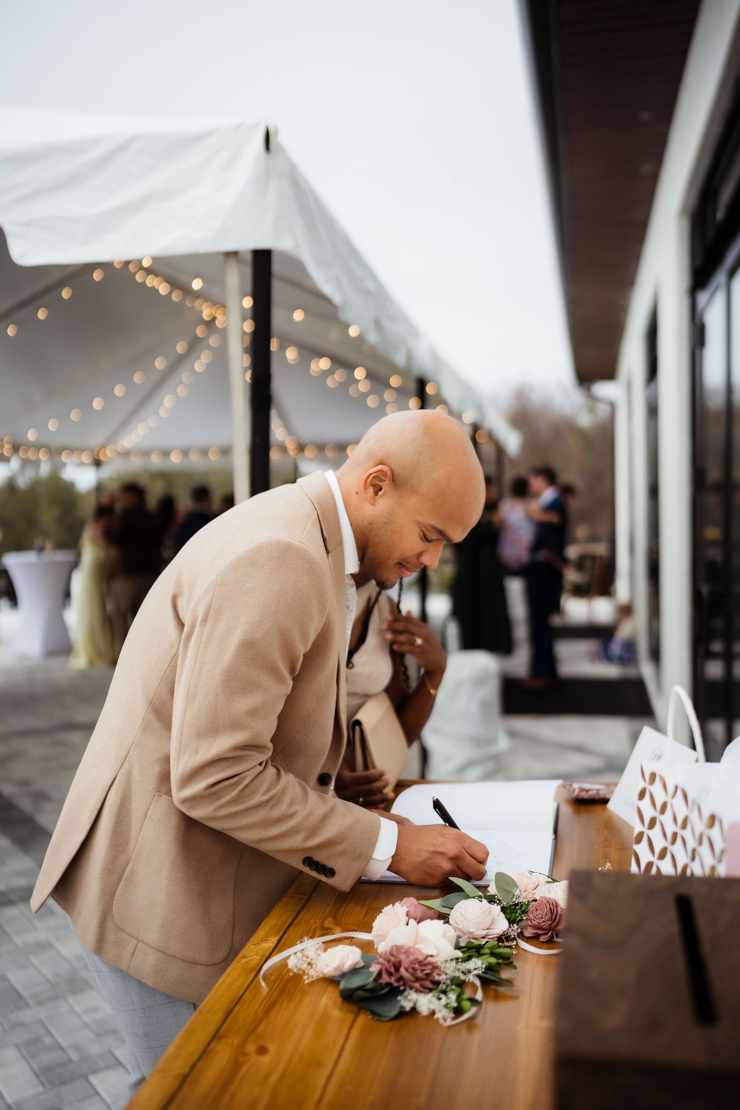 Person in beige blazer signing documents at an outdoor venue with string lights and floral decorations.