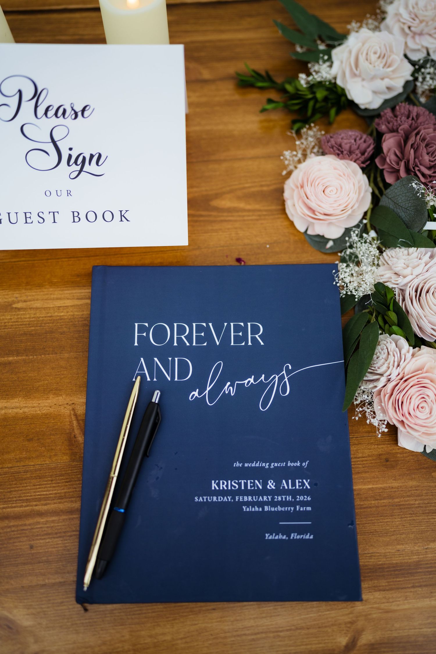 Navy blue wedding guest books and signage with elegant calligraphy displayed alongside blush roses on wooden surface.
