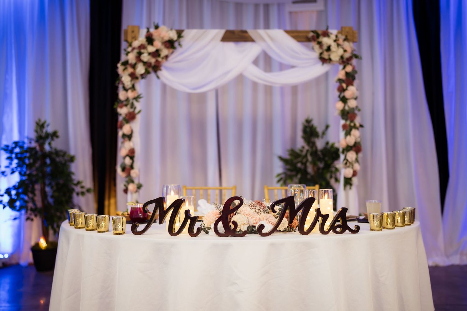 White fabric drapes and floral arrangements create an elegant wedding ceremony backdrop with blue accent lighting.