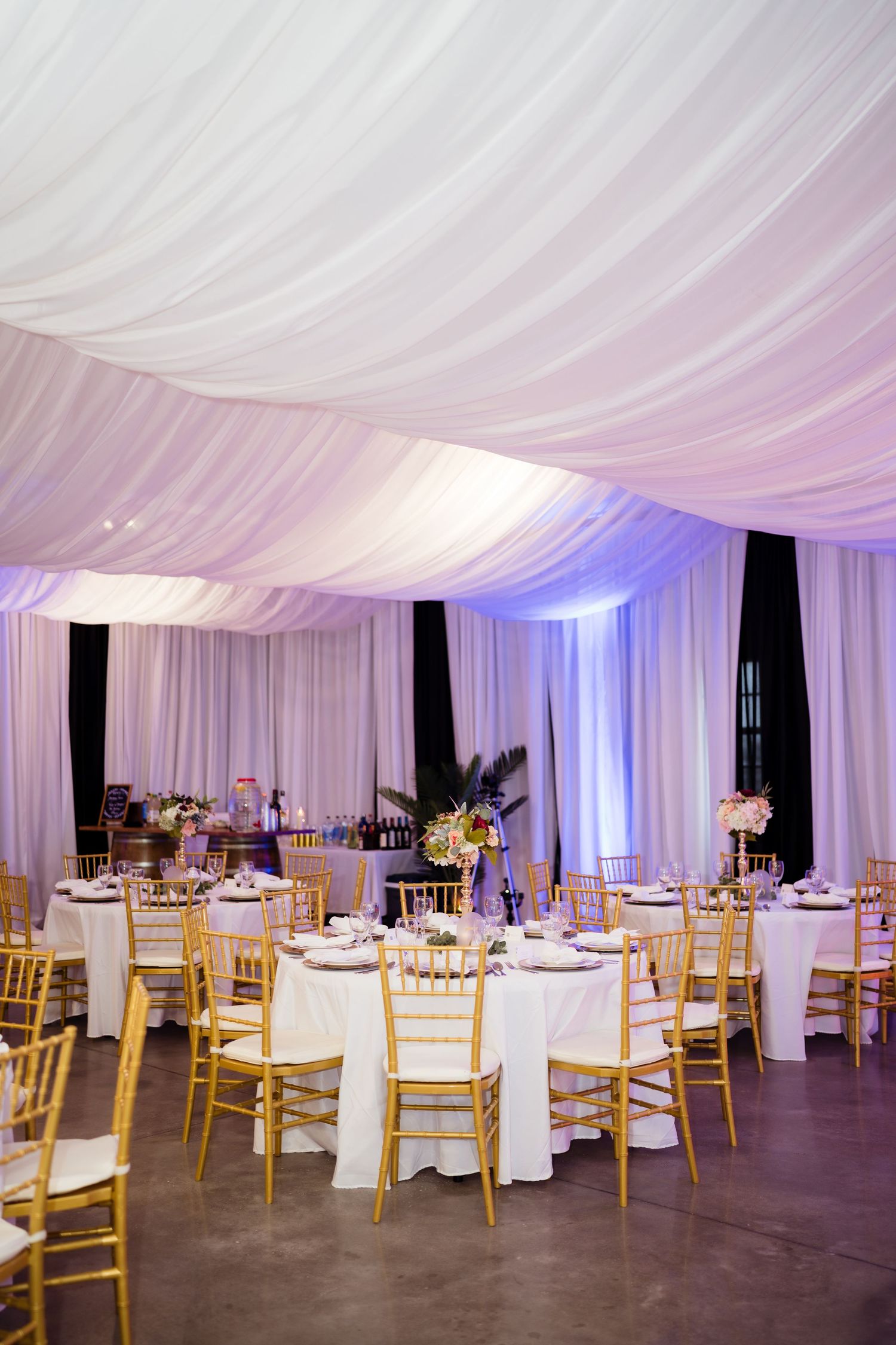 Elegant wedding reception venue with white draped ceiling, blue uplighting, and gold chiavari chairs around round tables.