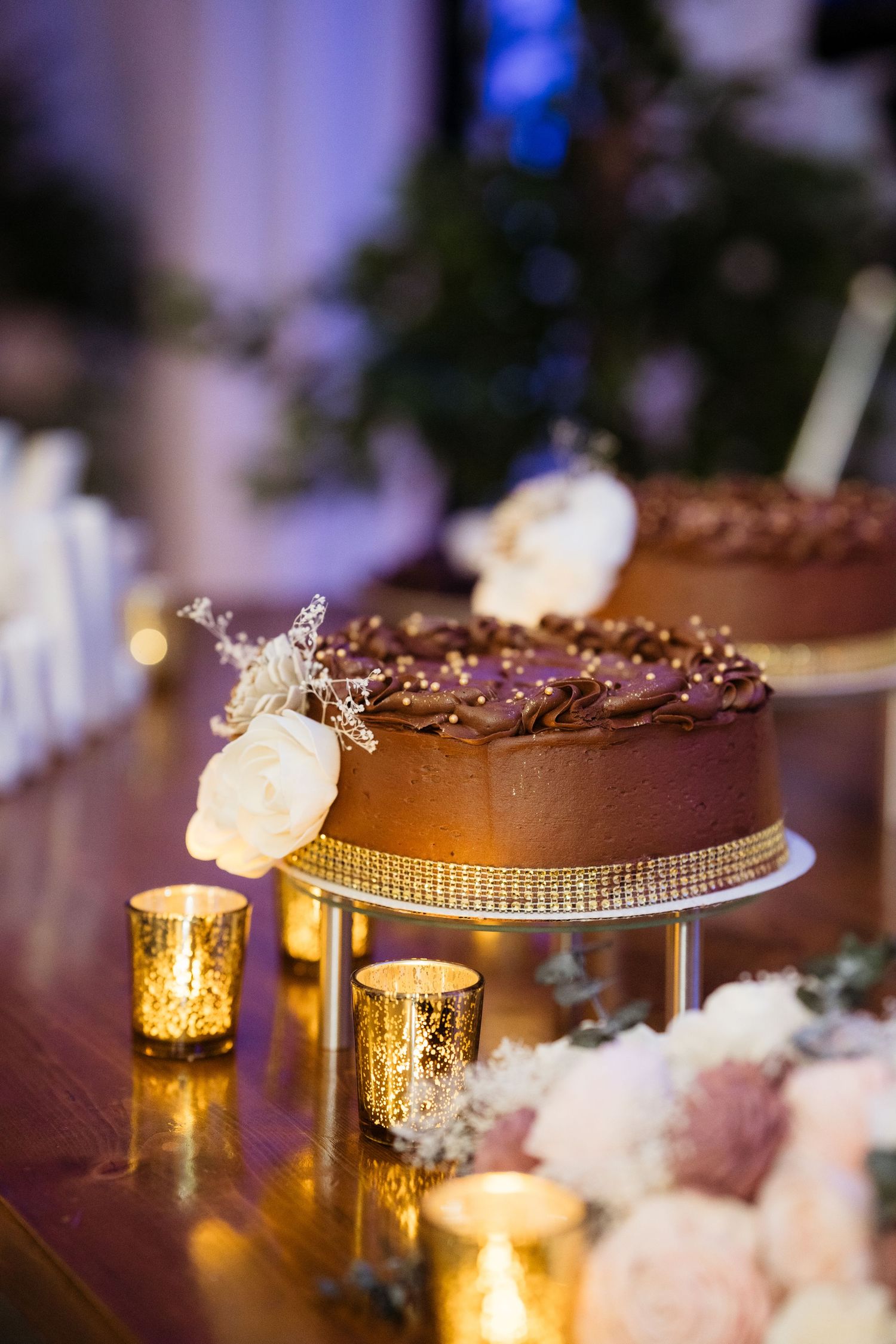 Elegant chocolate wedding cake displayed on gold stand with glowing candles and cotton decor creating romantic ambiance.
