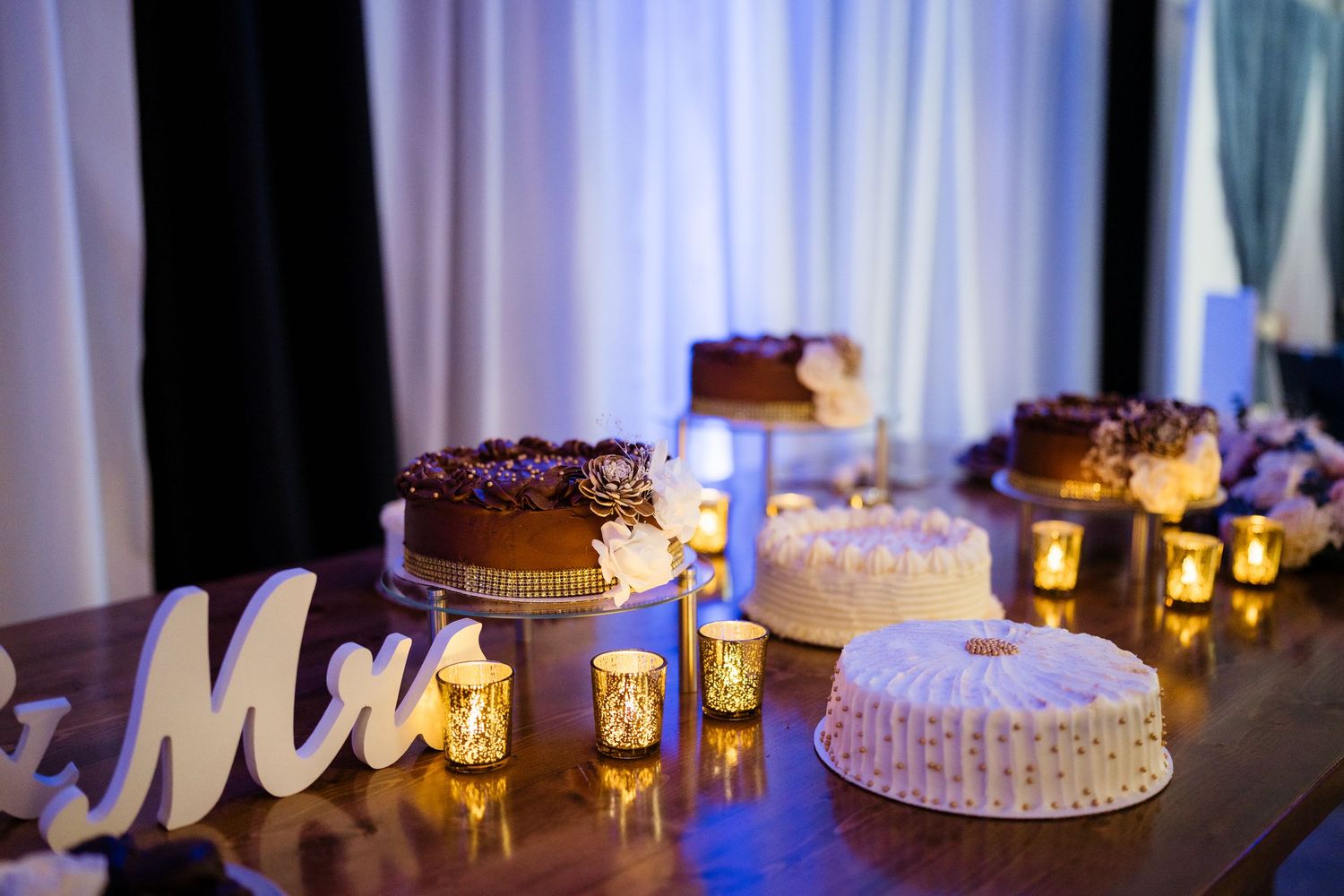 A dessert table with illuminated candles displays multiple cakes and a decorative 'Mr' sign at an elegant event.