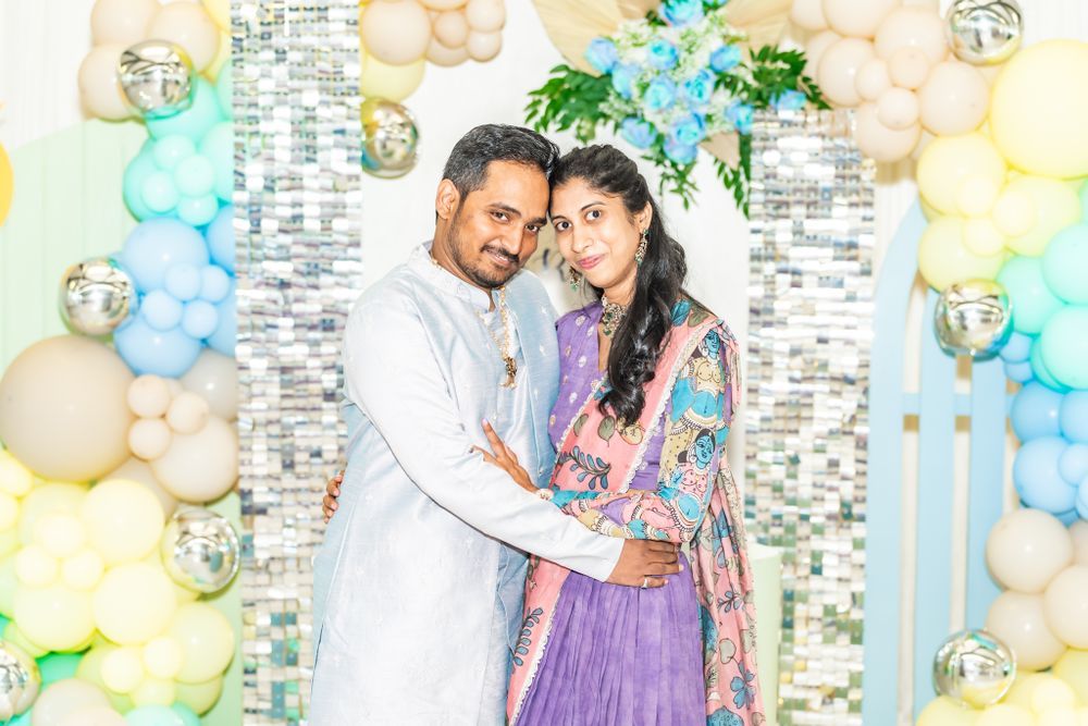 A couple poses together at a celebration with pastel balloon decorations and floral arrangements in the background.