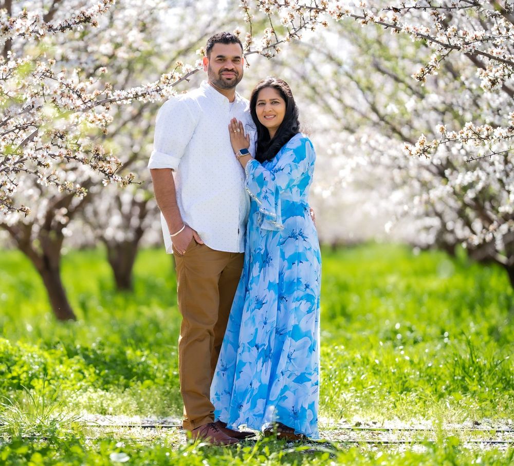 A couple poses together in a spring orchard with blooming white cherry blossoms and green grass.