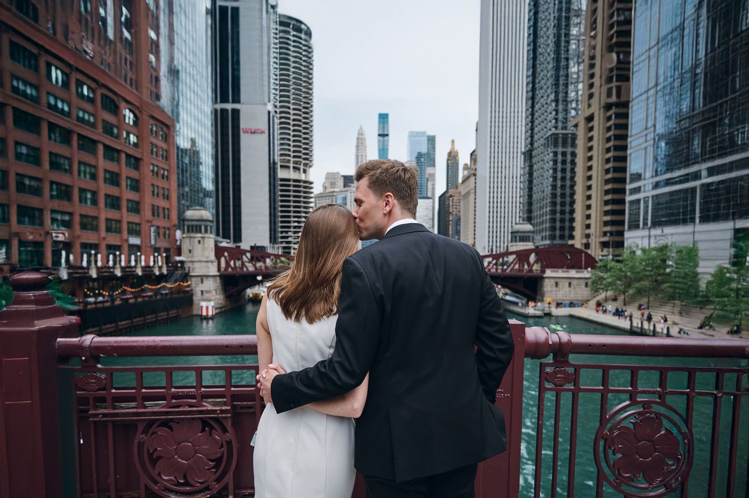 A couple in wedding attire embraces on a bridge overlooking a city river with tall buildings in the background.