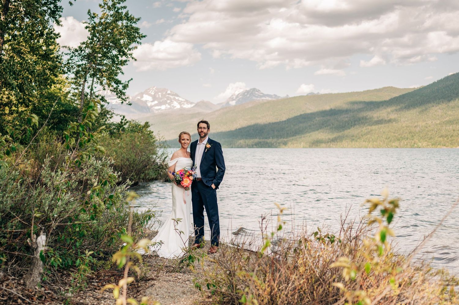 Couple celebrates their wedding day against a scenic mountain lake backdrop with cloudy skies and natural foliage.