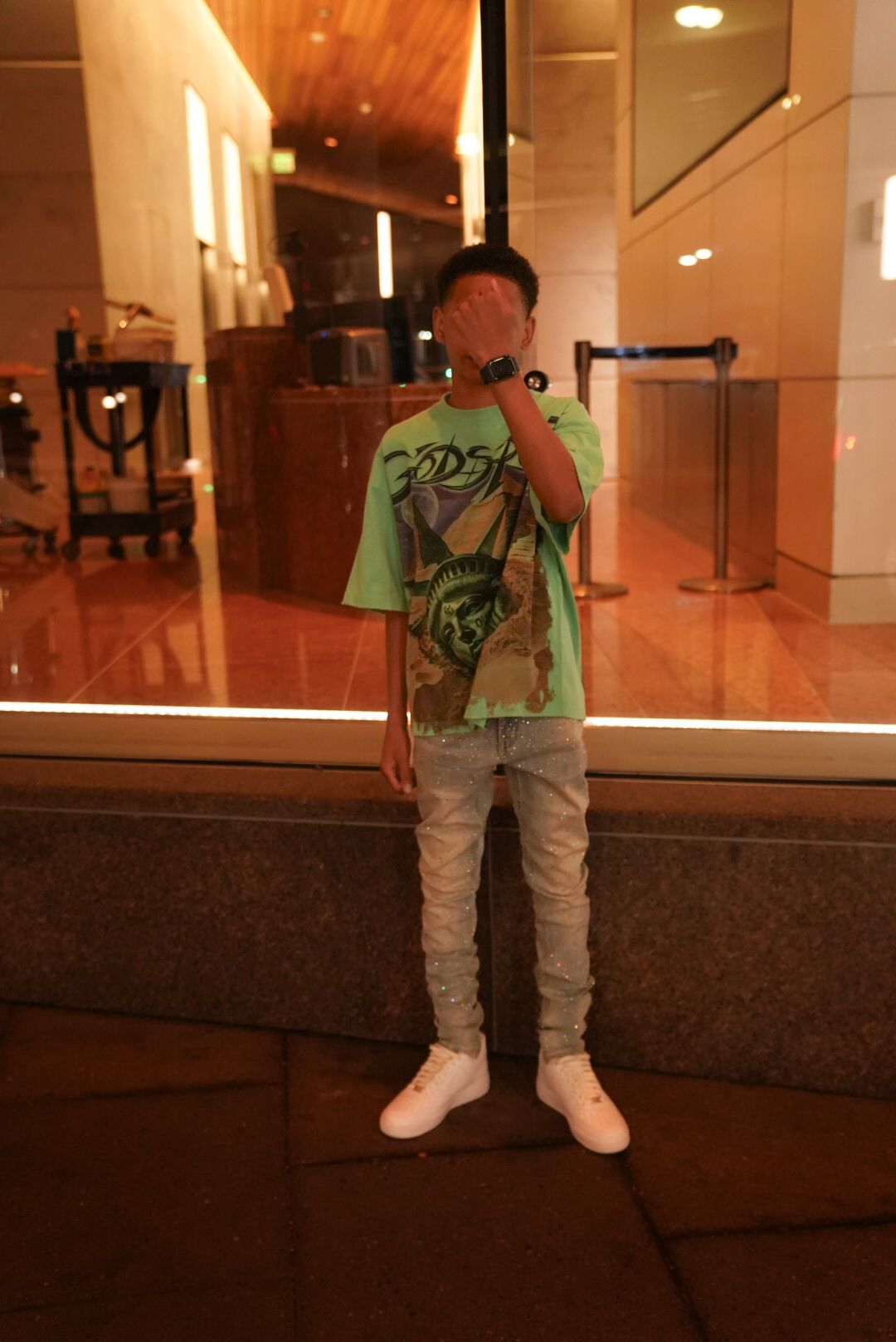 Person wearing a bright green t-shirt and white pants stands in front of an illuminated indoor space at night.