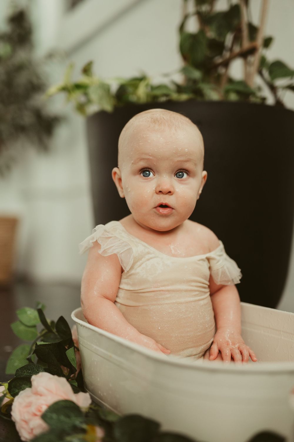 A cute baby in a cream romper sits in a small white tub surrounded by pink roses and green plants.
