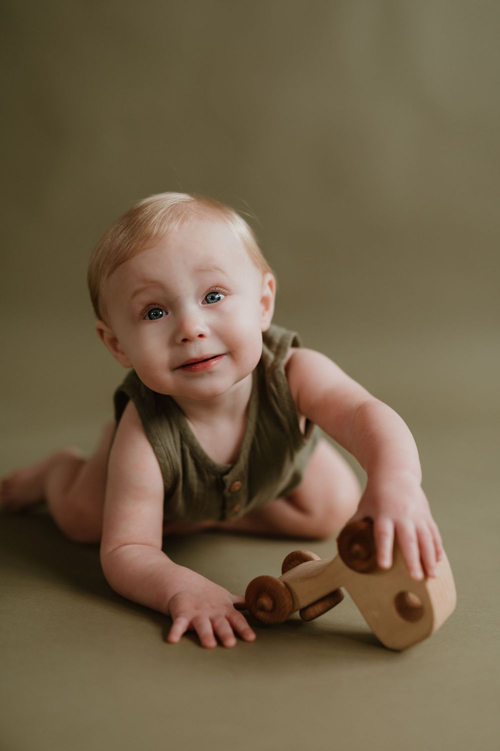 Smiling baby in olive romper plays with wooden toy on matching green background.