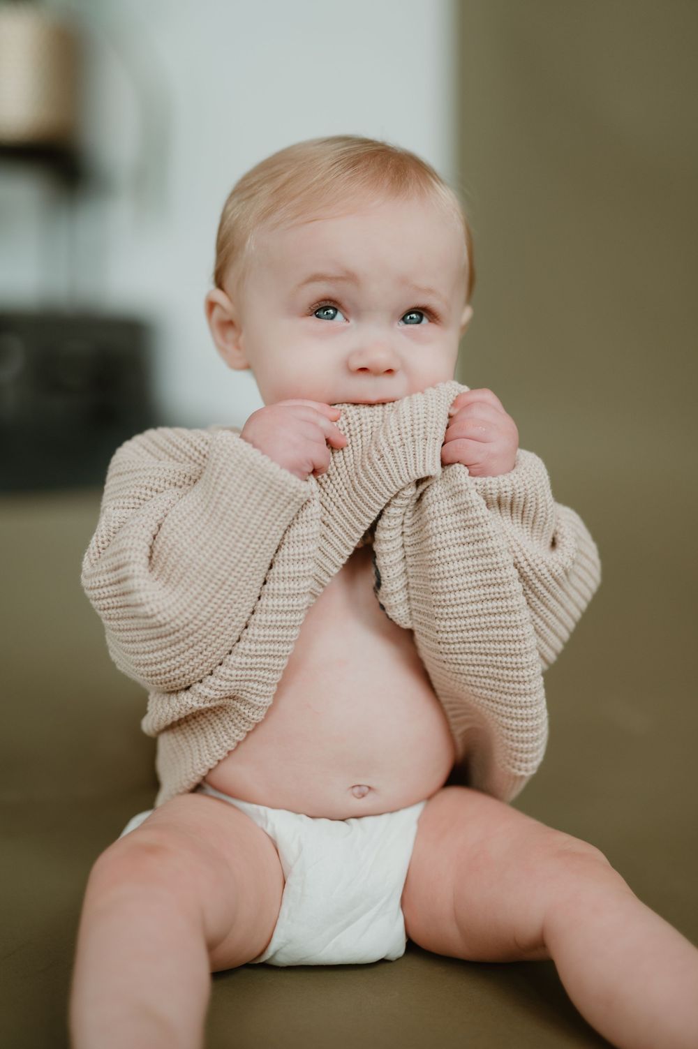 Adorable baby in a beige knit sweater and diaper, chewing on shirt hem with bright blue eyes.