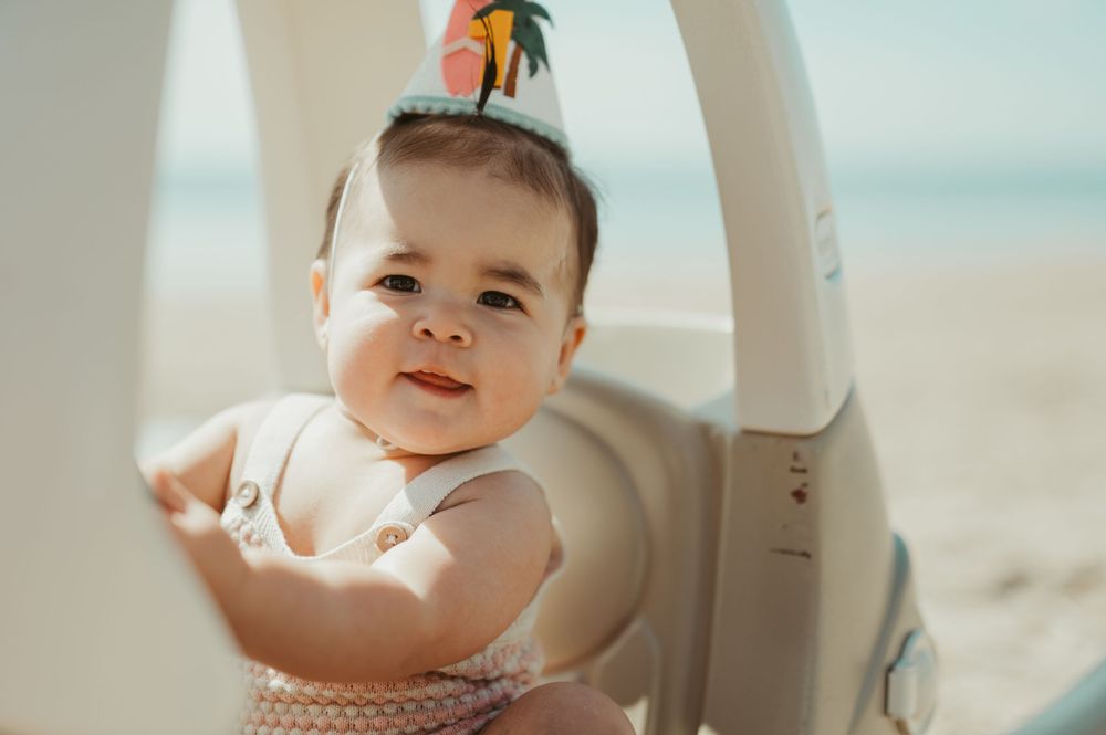 Adorable baby wearing a party hat smiles while sitting in a white chair on a sunny beach.